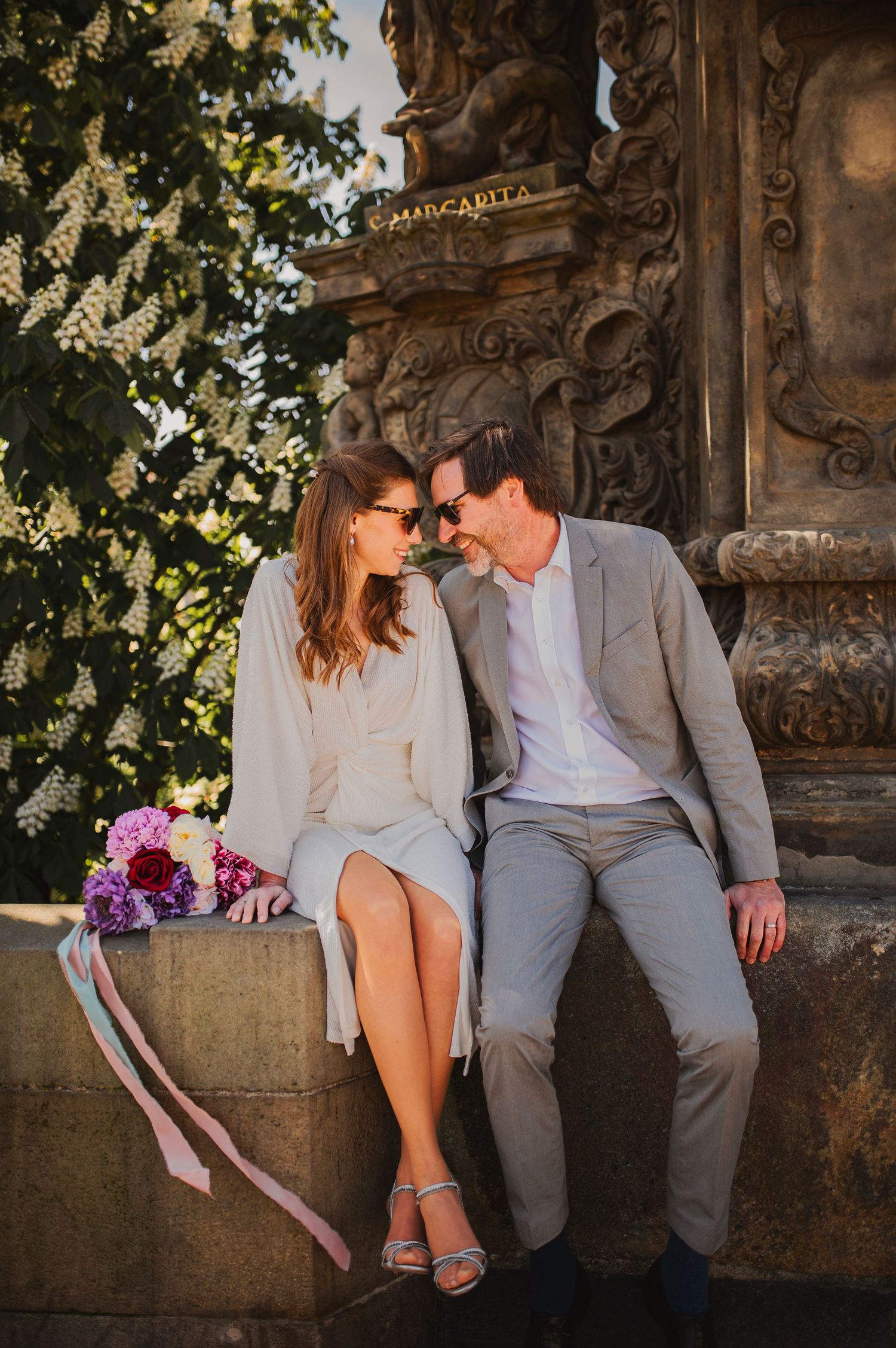 Couple shares tender moment on ornate stone ledge with purple bouquet at golden hour.