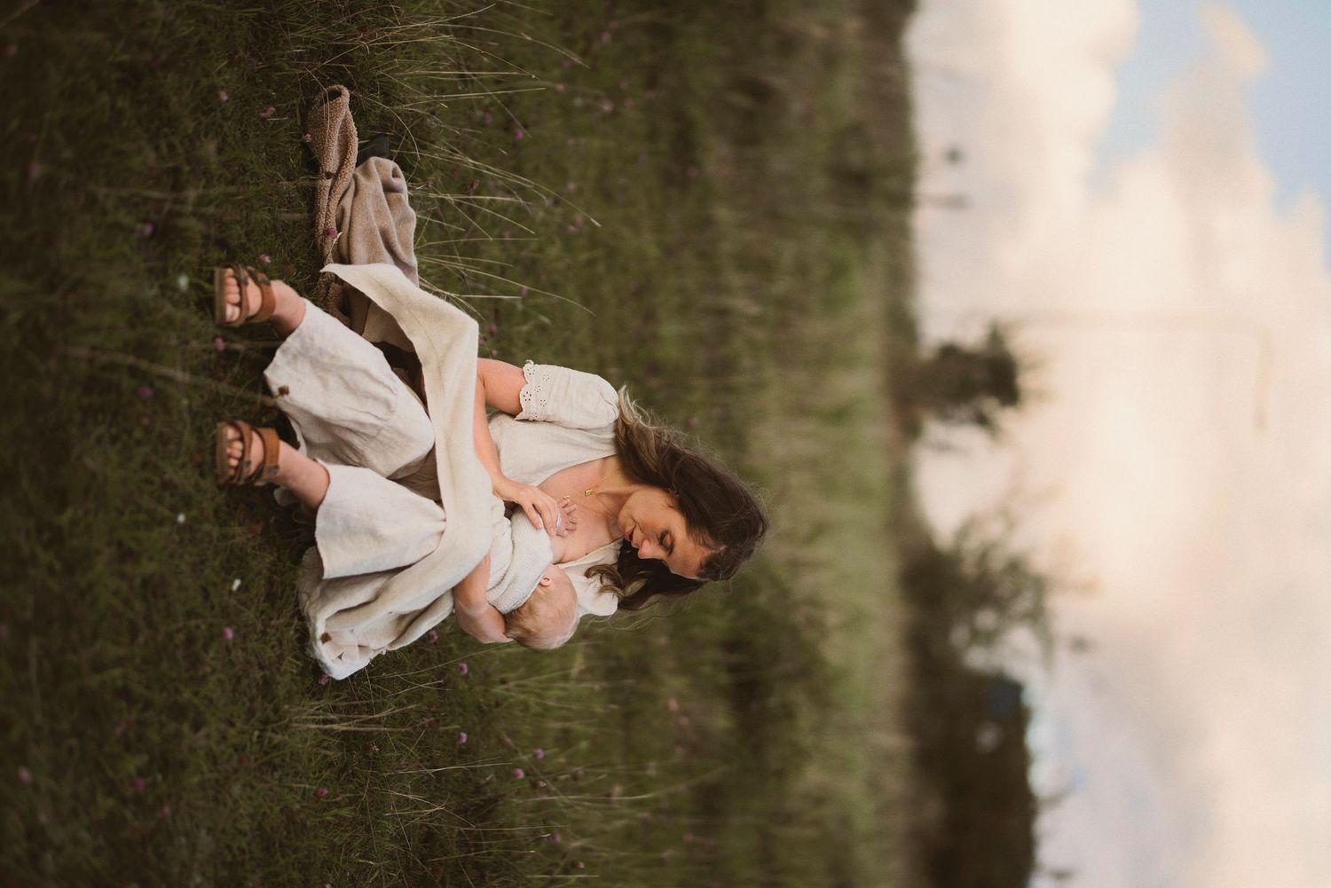 Parents cuddling with baby in white outfits on grassy hill at sunset.