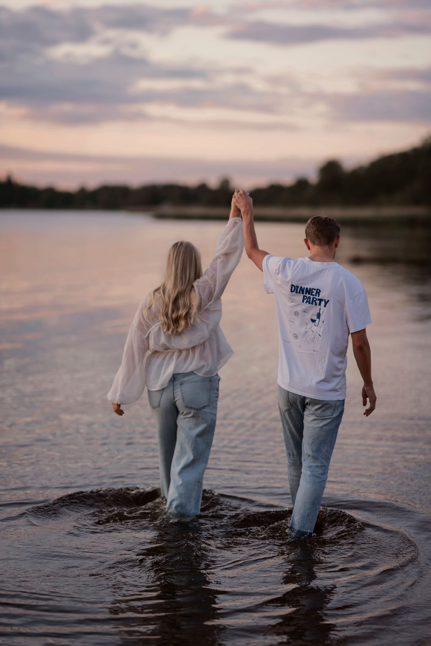 A couple holds hands while wading in water at sunset with cloudy skies above.