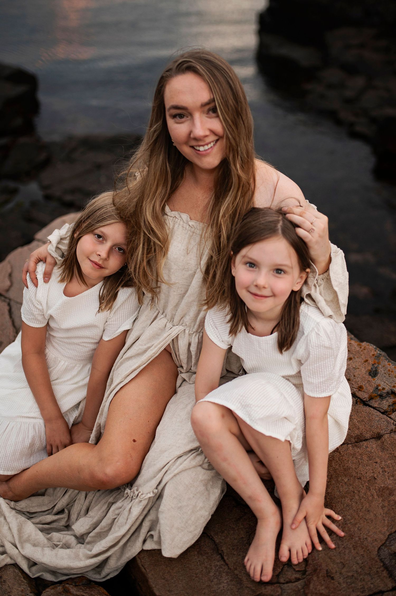 Family group in white and cream dresses sits together on coastal rocks at sunset.