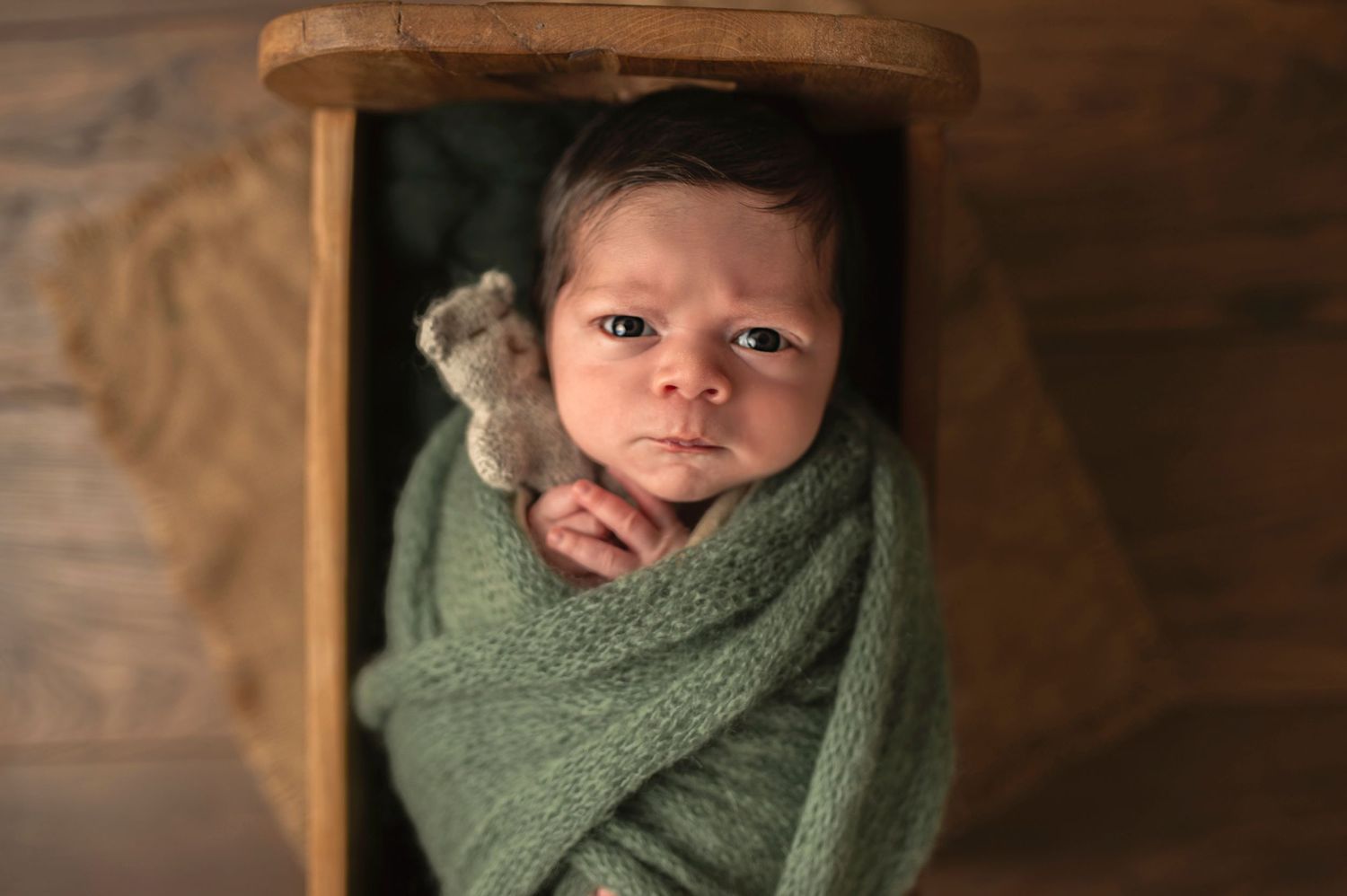 Newborn wrapped in soft sage green blanket rests in wooden box during professional photoshoot.