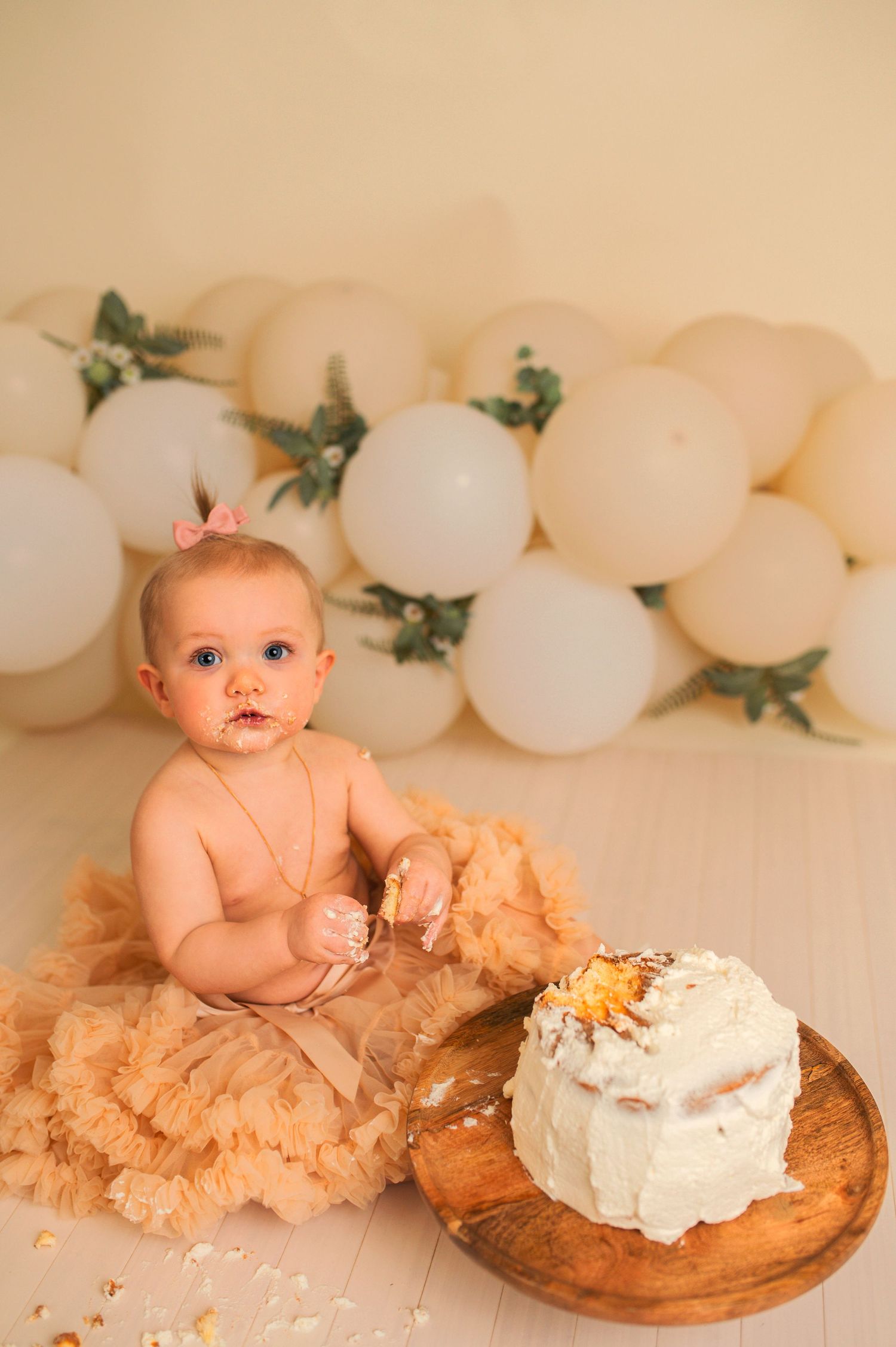 Birthday cake smash scene with white balloon arch and rustic wooden cake platform.