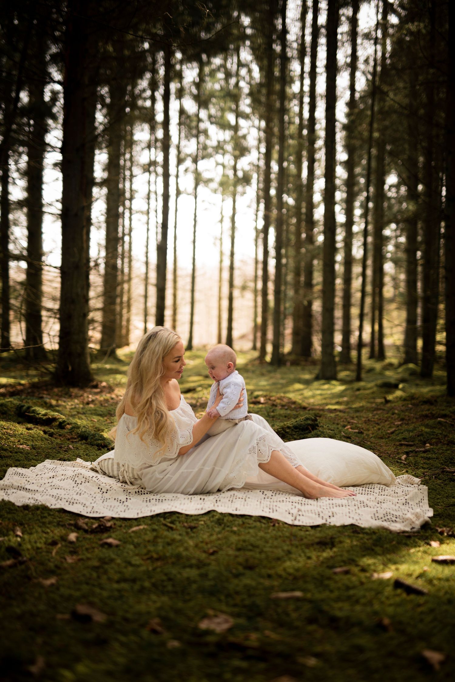 A tender moment captured in a sunlit forest with a cream blanket on mossy ground.