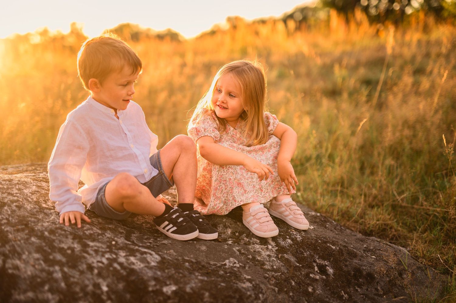 Two people sitting together on rock in sunny field during sunset.