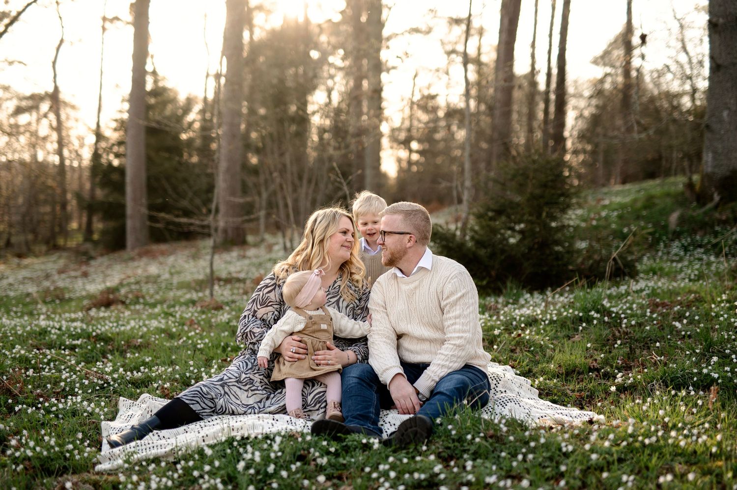 Family of four sitting on a blanket in a field of white flowers surrounded by trees during golden hour.