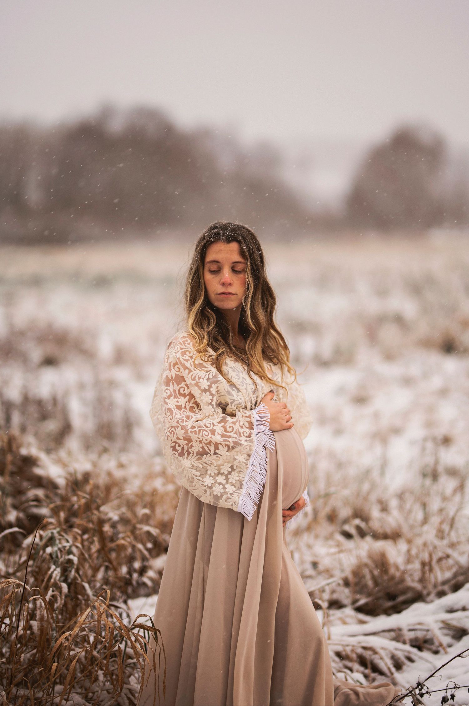 A pregnant woman in a beige lace dress poses thoughtfully in a snowy winter field.