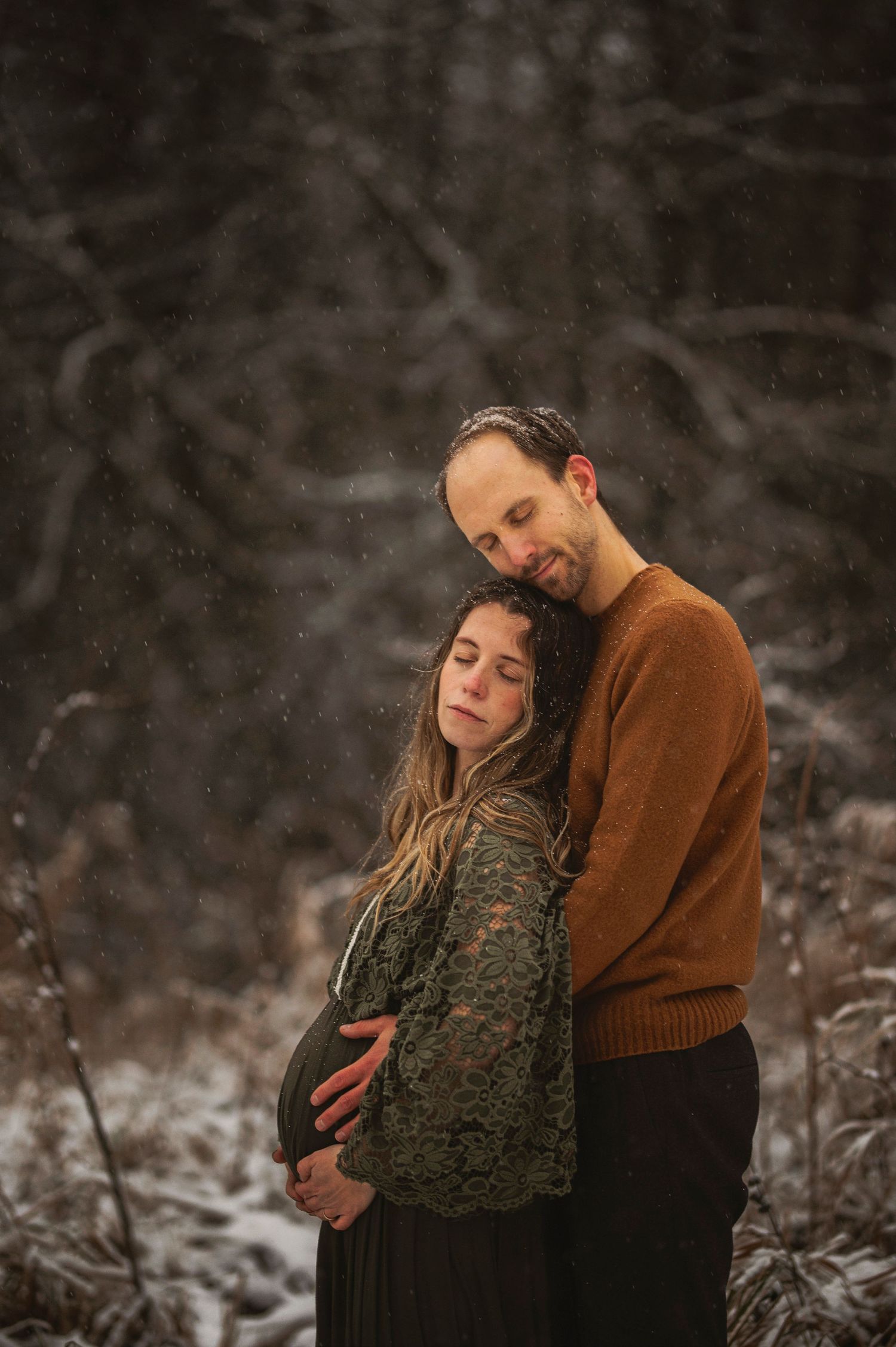 Intimate winter maternity portrait showing couple embracing in rustic snowy forest setting.