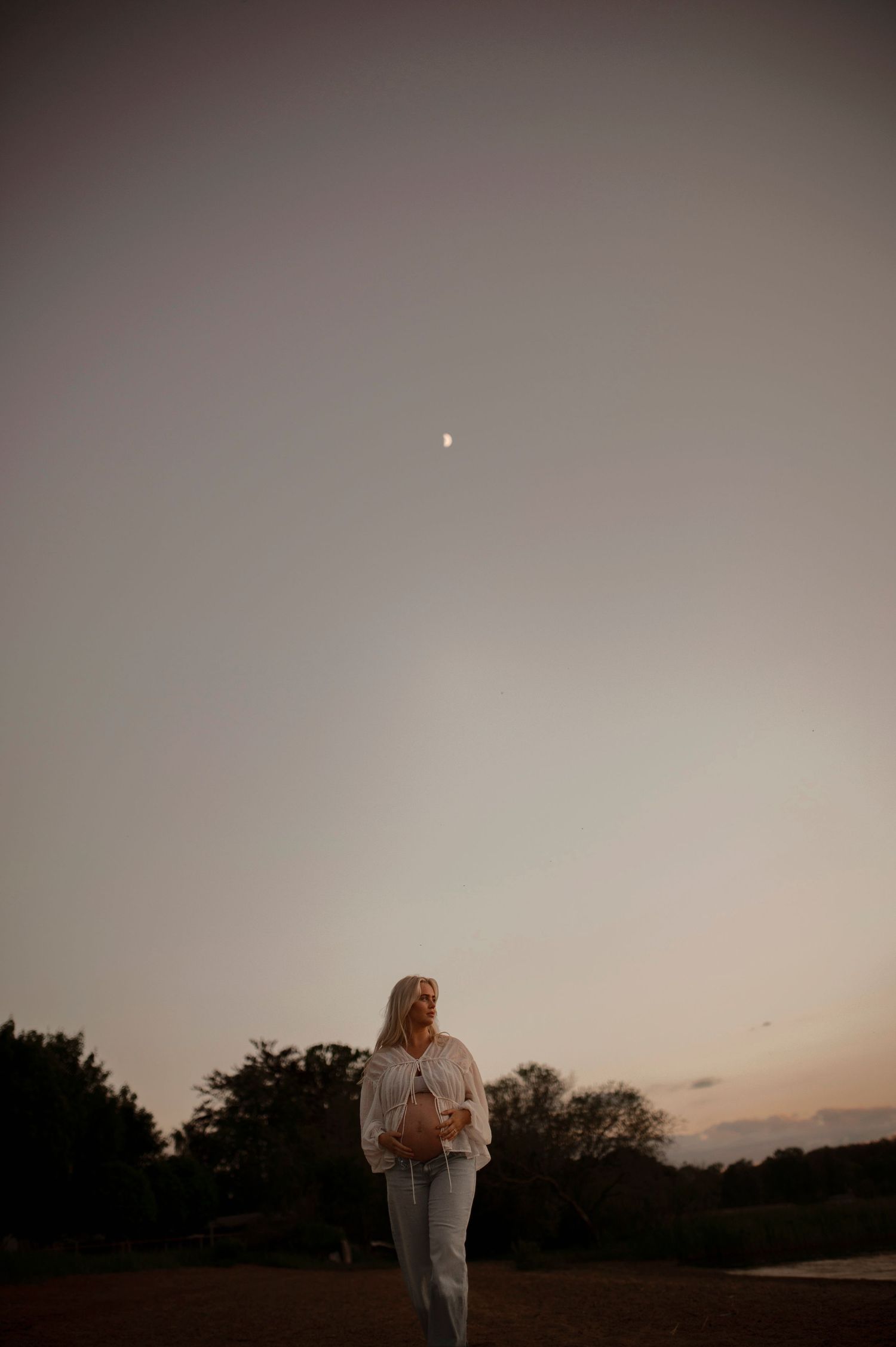 A silhouette of a person stands in a serene field at dusk with a crescent moon visible in the hazy evening sky.
