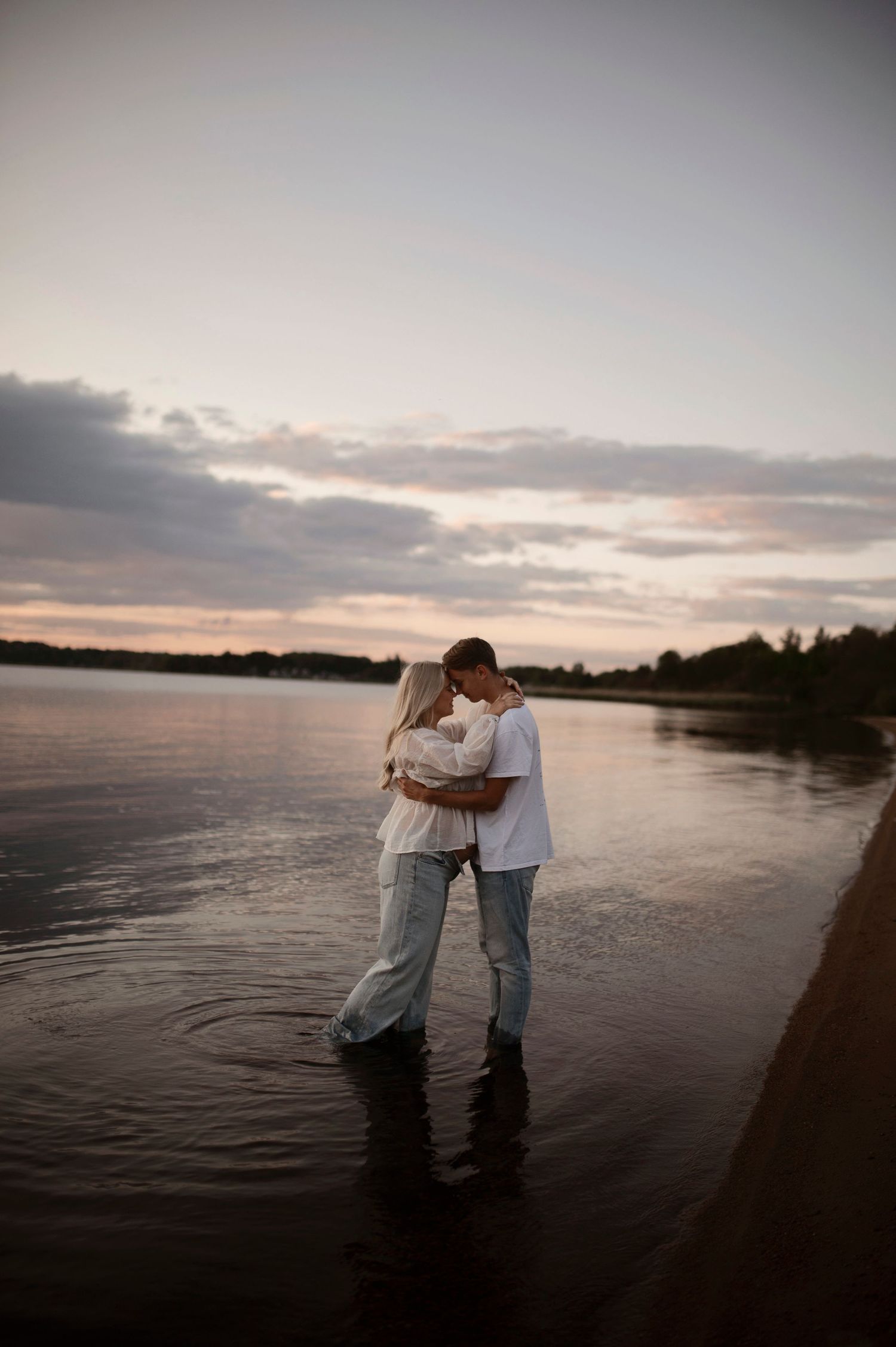A couple standing in shallow water during sunset, sharing an intimate moment by the lakeside.
