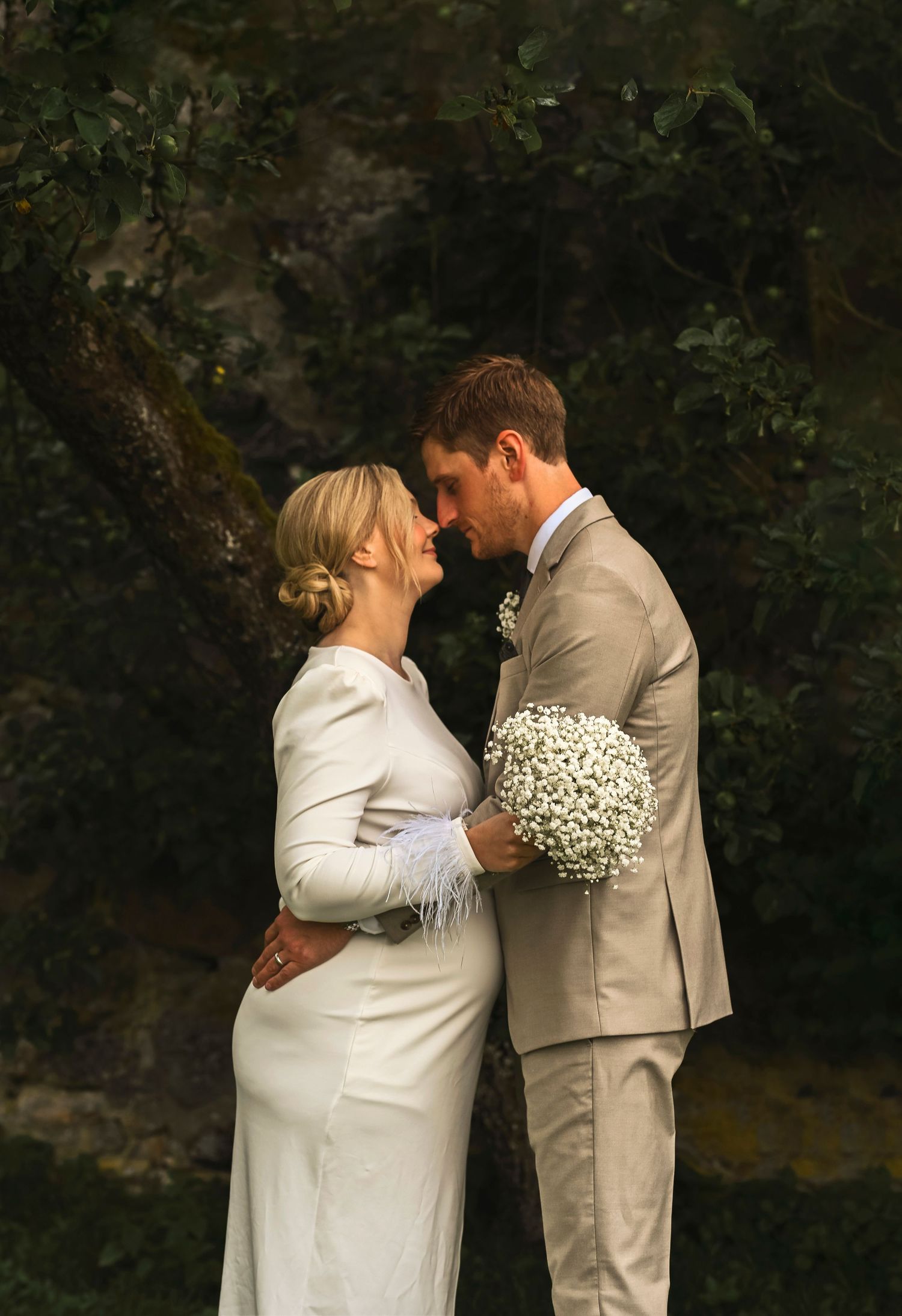 A wedding couple shares an intimate moment with baby's breath bouquet against a dark natural backdrop.