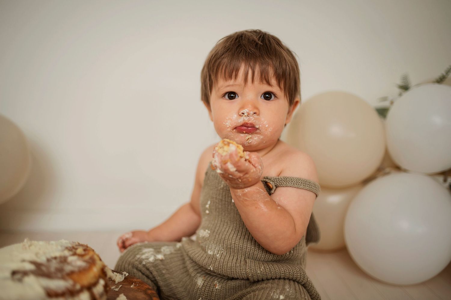 Baby in a beige knit romper sits on floor with white balloons and birthday cake during a cake smash photo session.