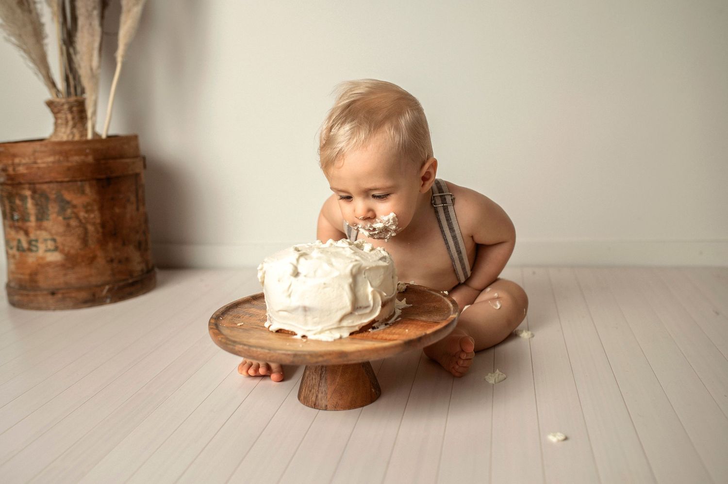 A baby in white sits on a wooden floor next to a rustic cake smash setup with vintage decorative elements.