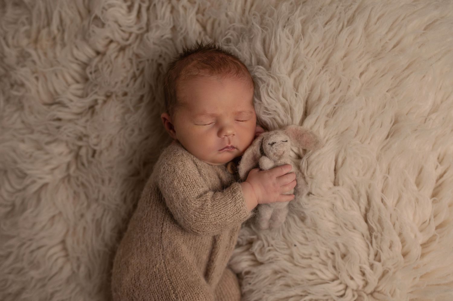 Sleeping newborn baby in beige knit outfit cuddling a soft bunny toy on a fluffy cream fur blanket.