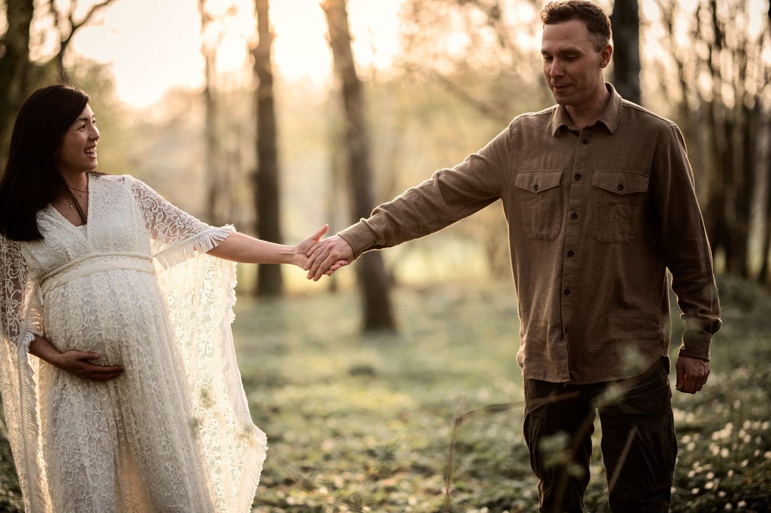 Couple holds hands while posing in white lace dress and brown jacket during forest photoshoot.