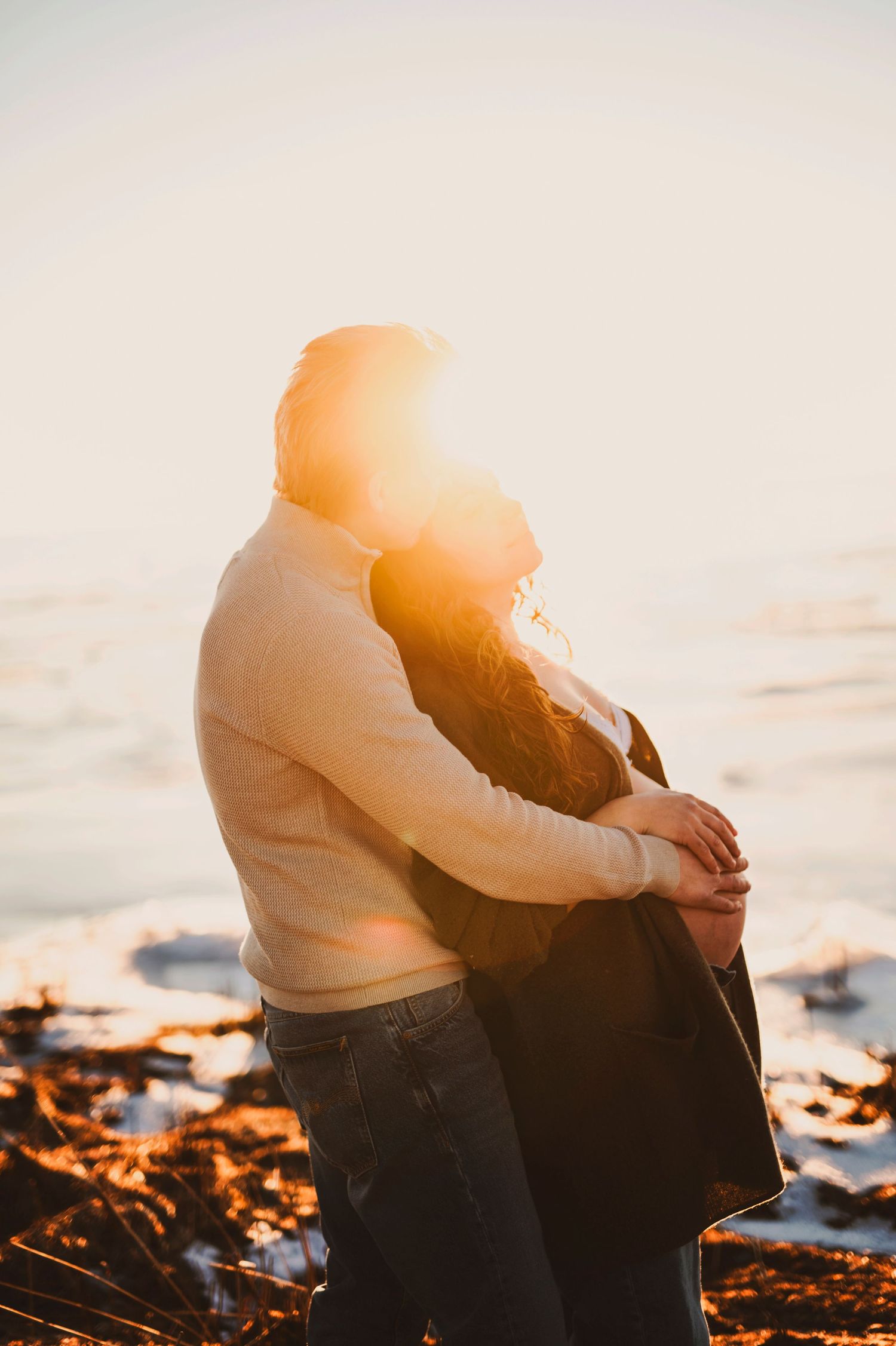 Couple embracing on a rocky beach at sunset with warm golden light illuminating their silhouettes.