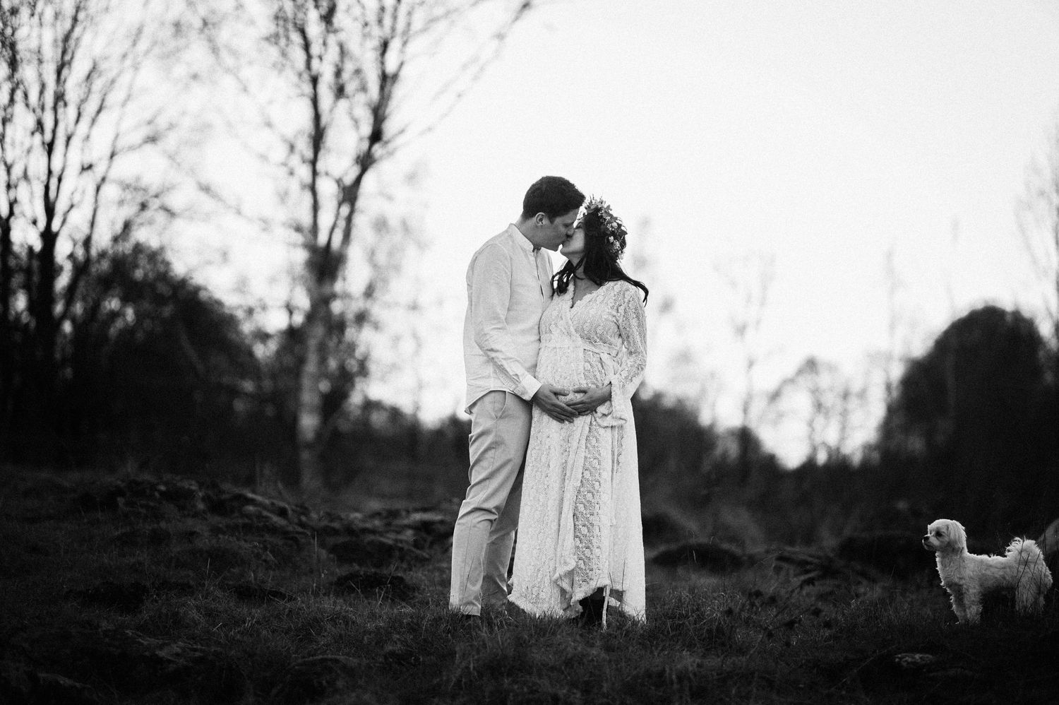 Black and white romantic photo of couple embracing on a hillside at dusk with a small dog in the background.