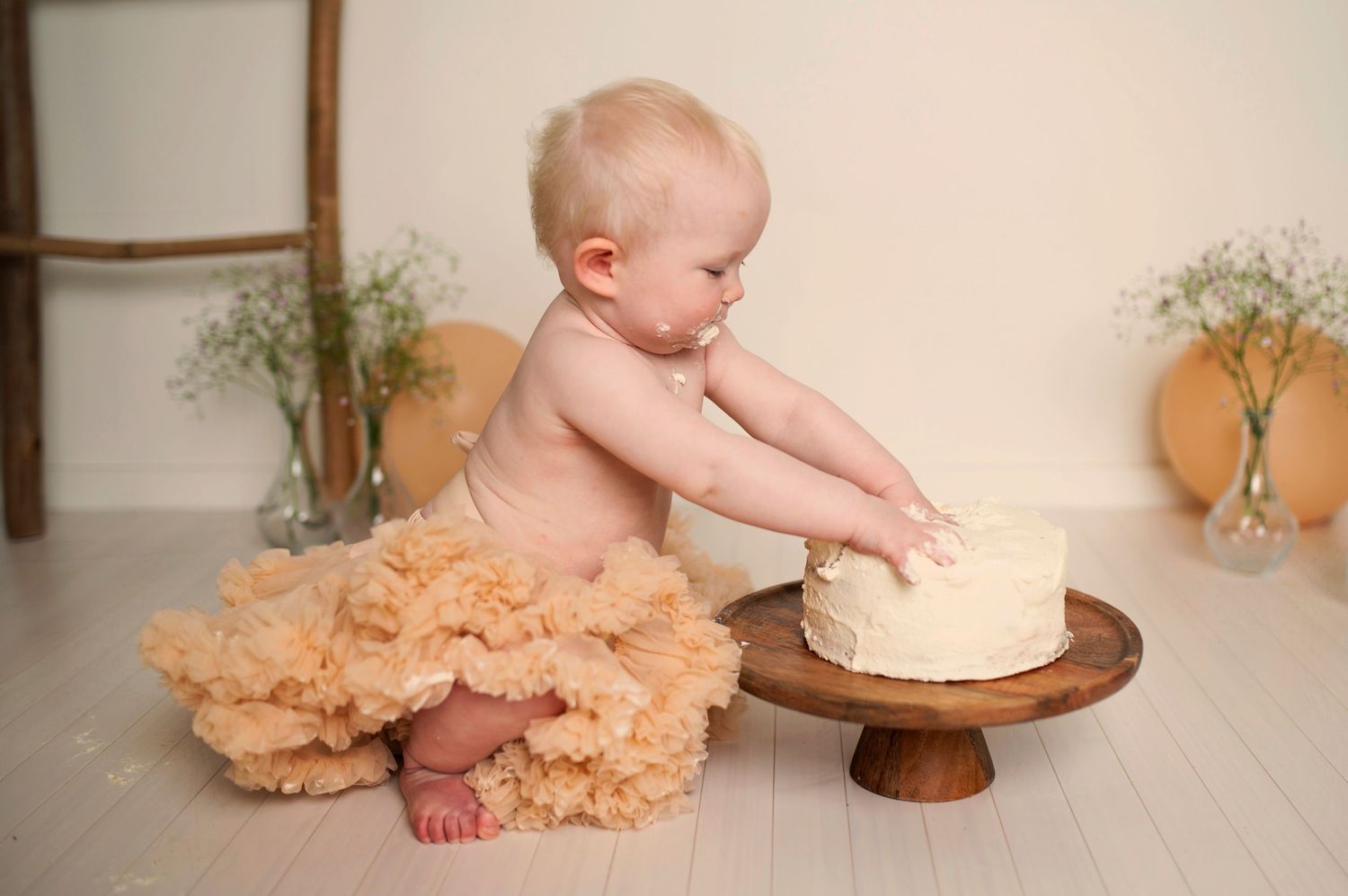Baby in peach tulle exploring white cake on wooden stand during first birthday photoshoot.