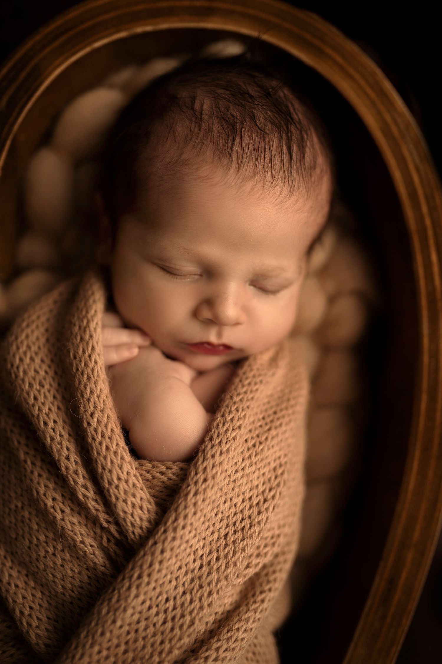 Sleeping newborn baby wrapped in tan knit blanket inside wooden frame, dark background with soft lighting.