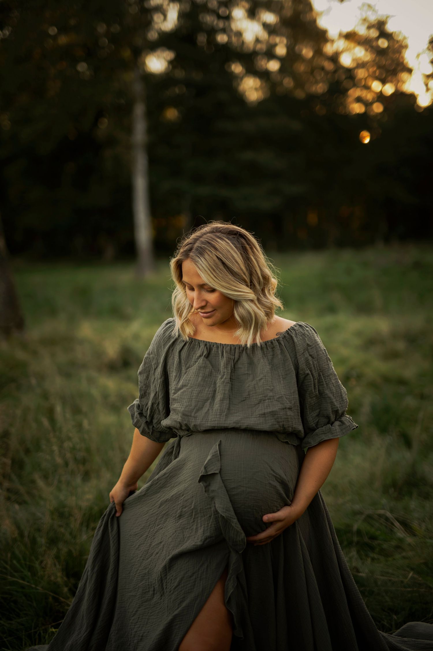 Side-view maternity portrait in gray dress against natural outdoor setting at dusk.