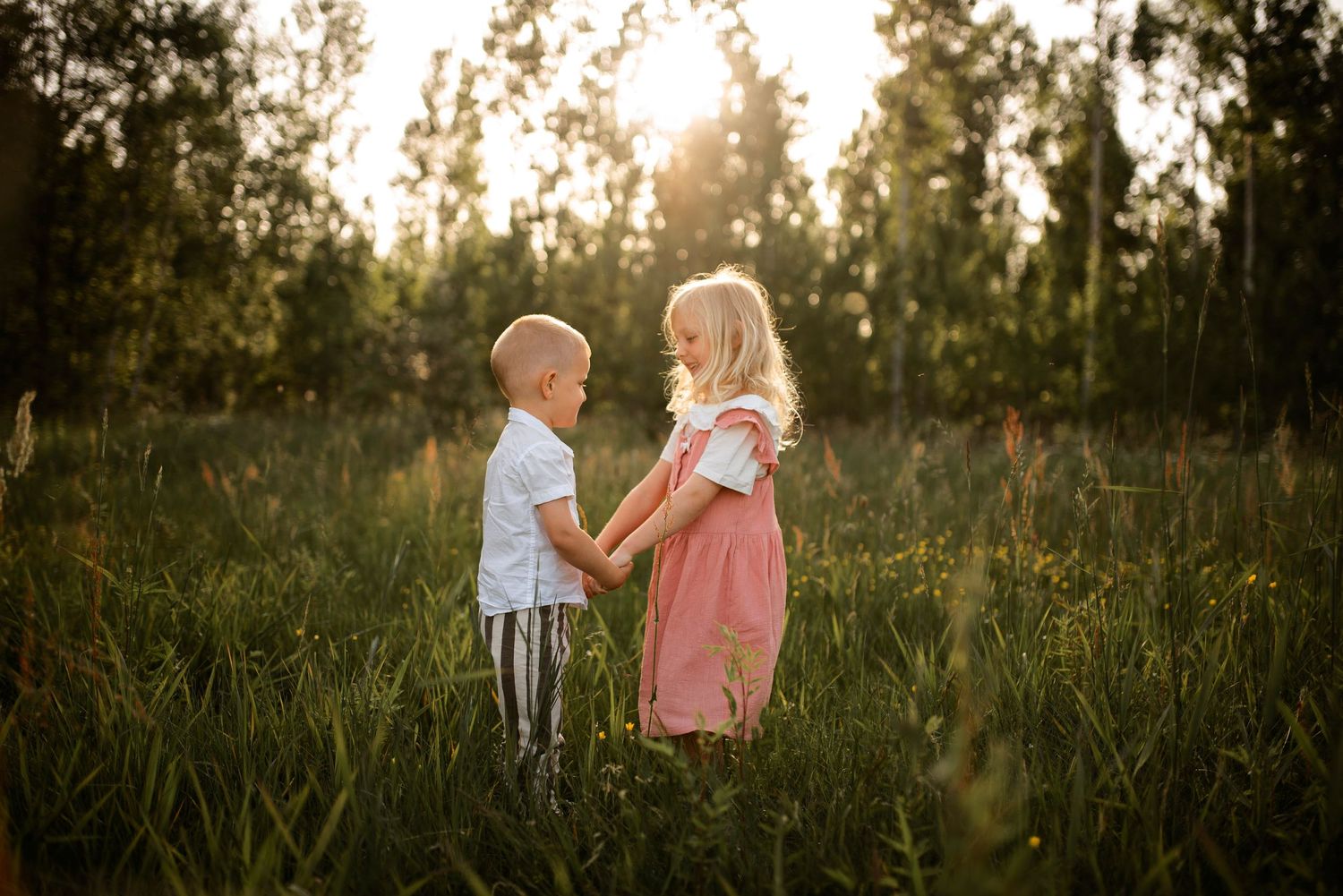 Two children standing together in tall grass at sunset.