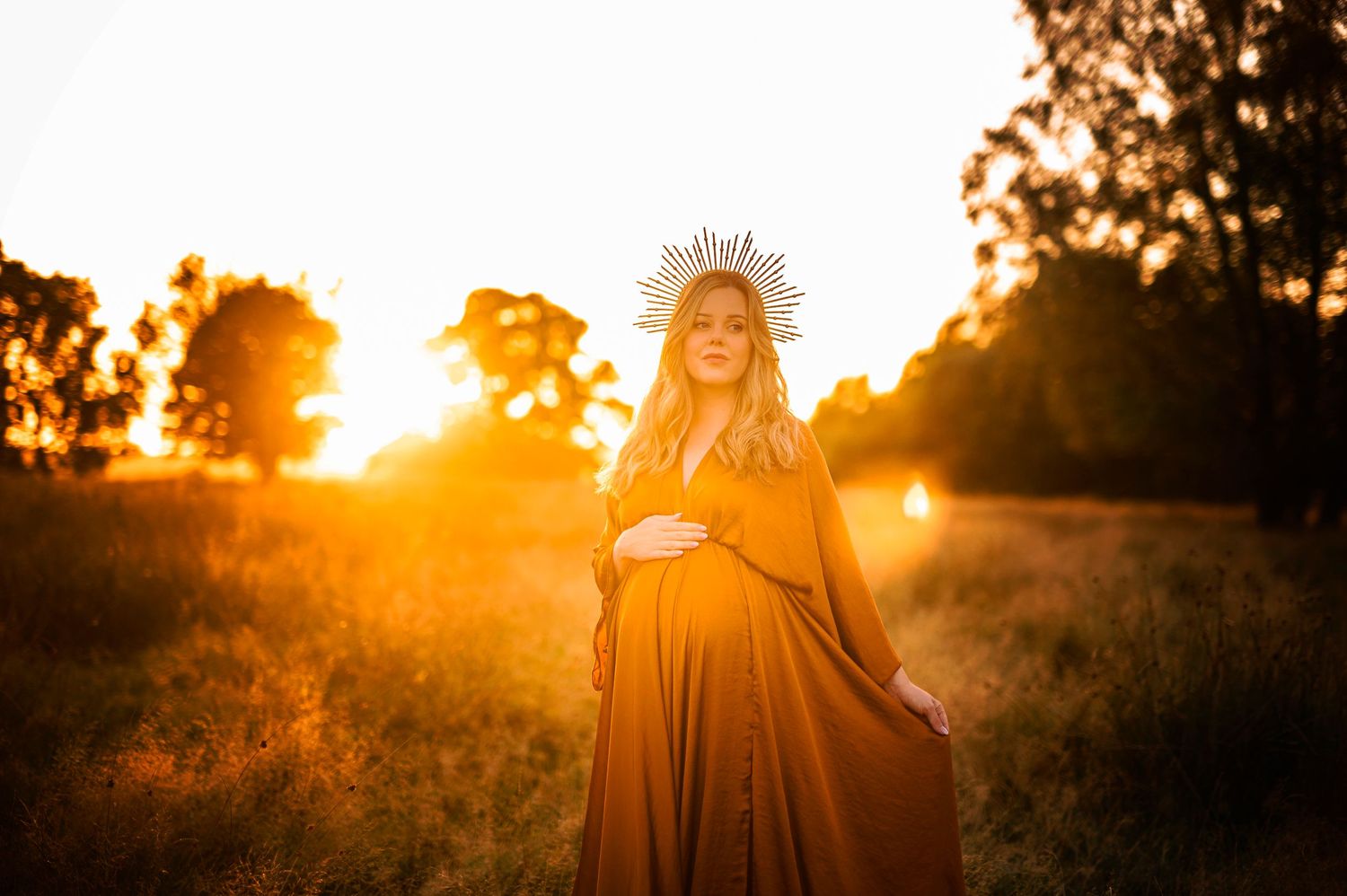 Glowing sunset maternity photo with sunburst crown effect in rustic outdoor setting.