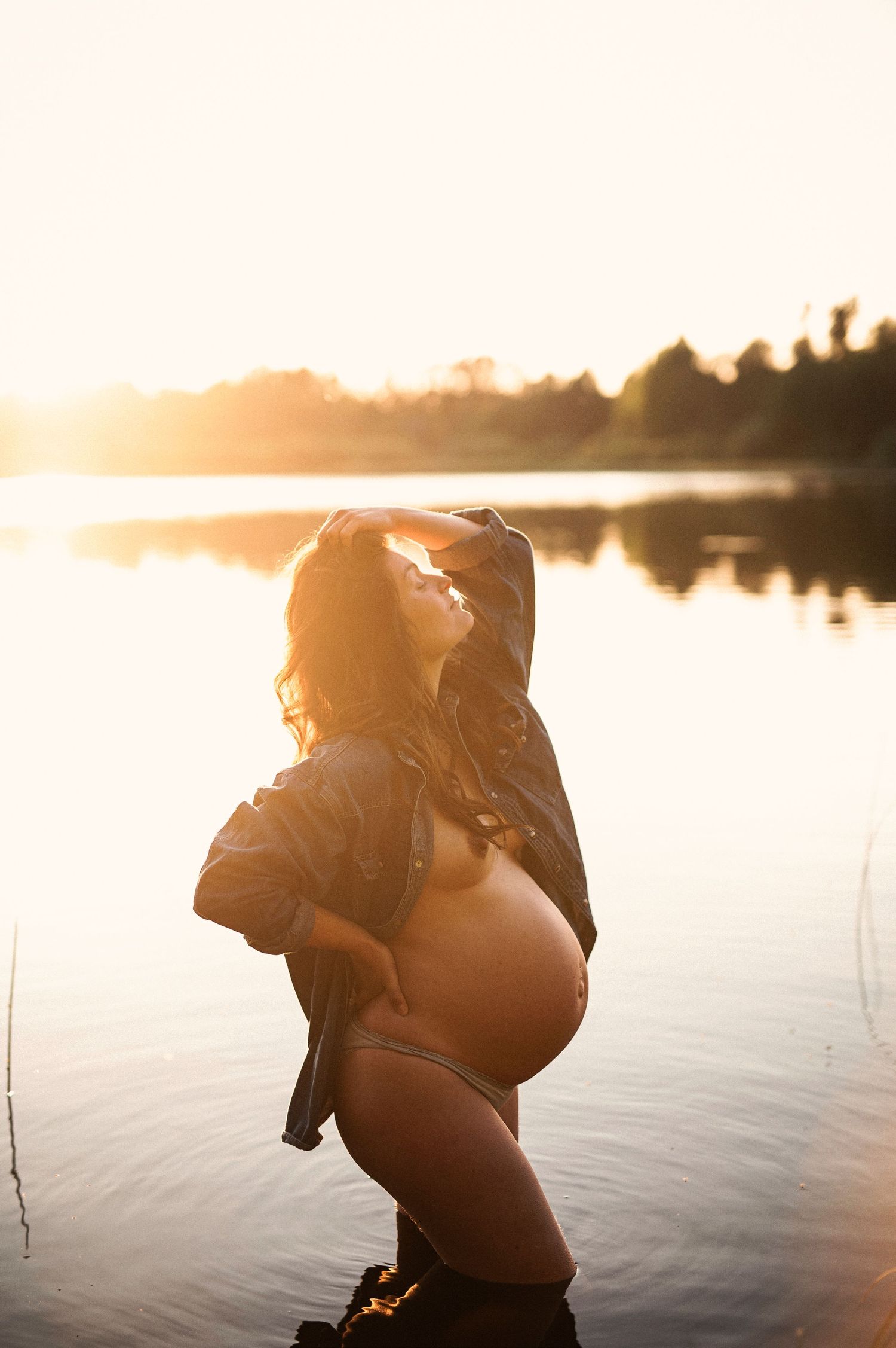 Pregnant woman standing in a lake at golden sunset, cradling her baby bump in warm glowing light.