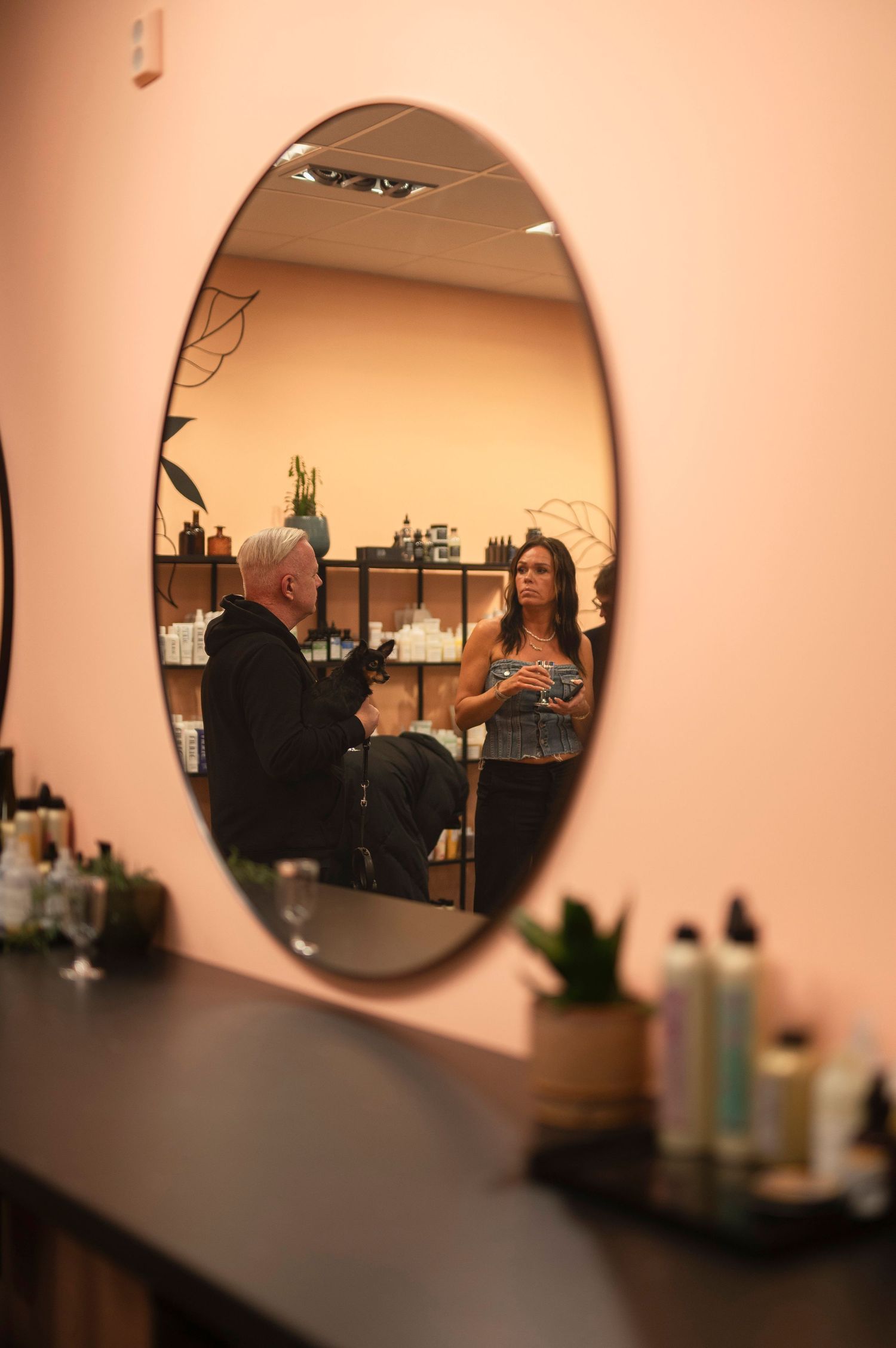 Hair stylist consulting with a client reflected in an oval mirror inside a pink salon.
