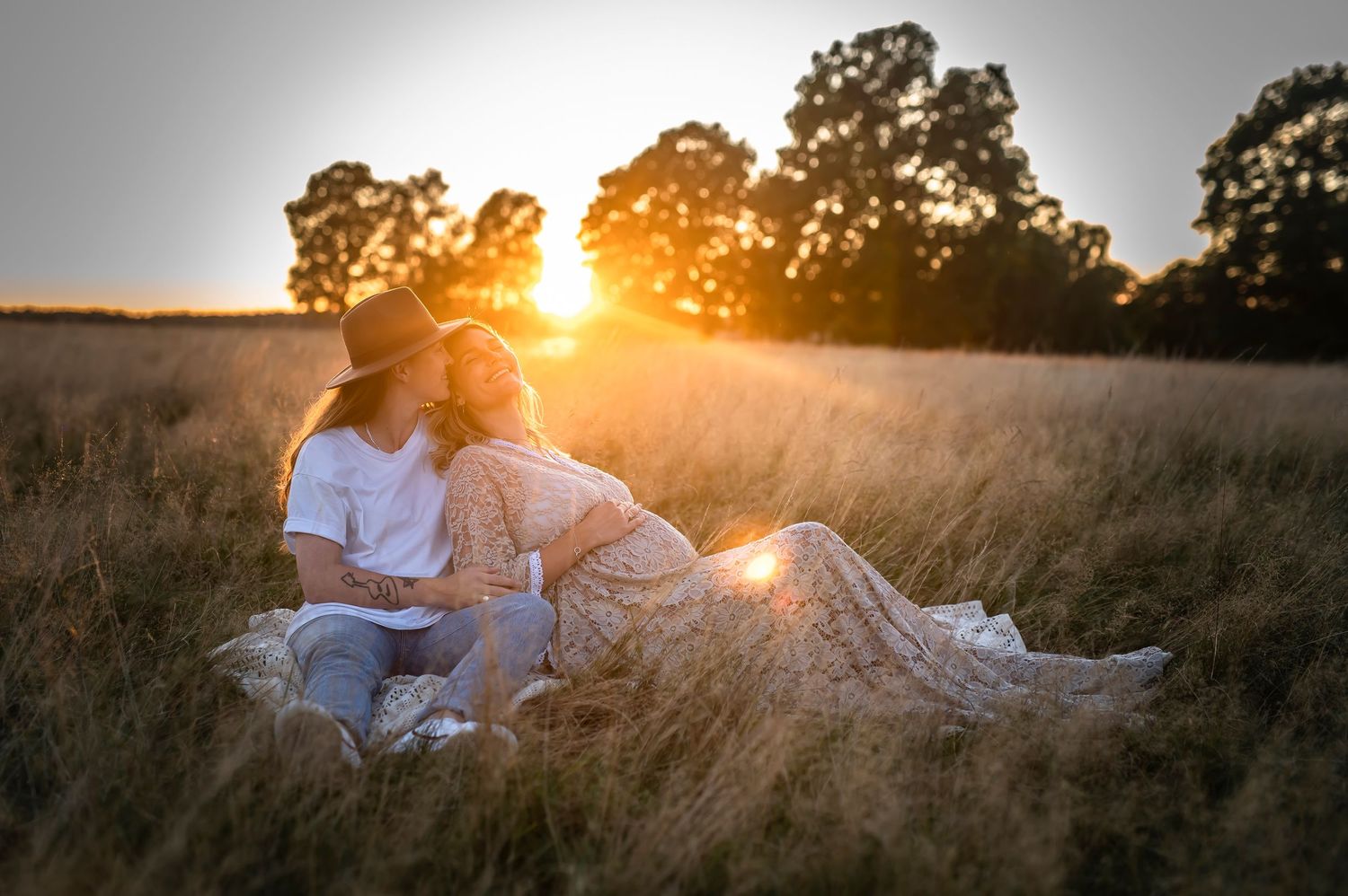 Couple sitting in golden field at sunset, pregnant woman in lace dress with partner in white tee, romantic maternity photo.