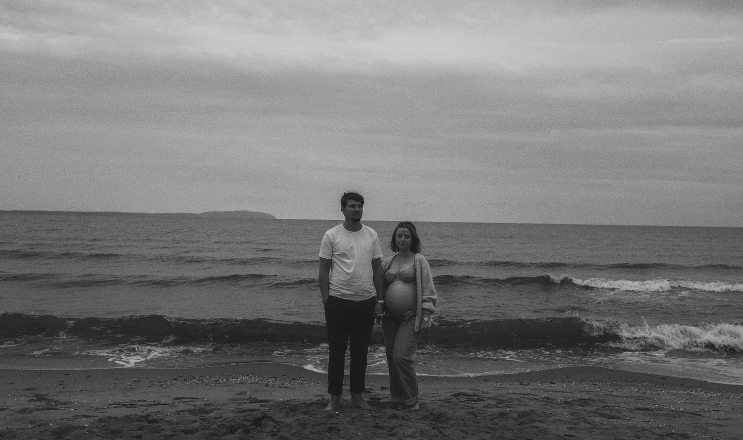 A couple stands together on a moody beach with waves crashing behind them under an overcast sky.