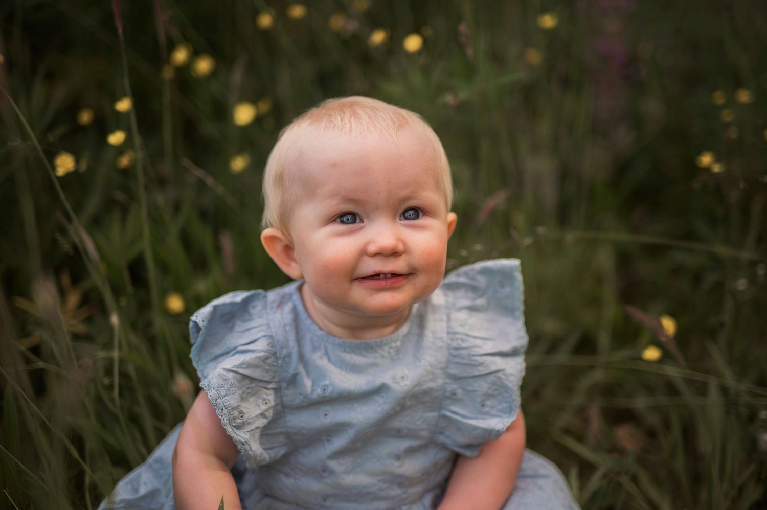 Smiling baby in ruffled blue dress poses in a field of wildflowers at sunset.