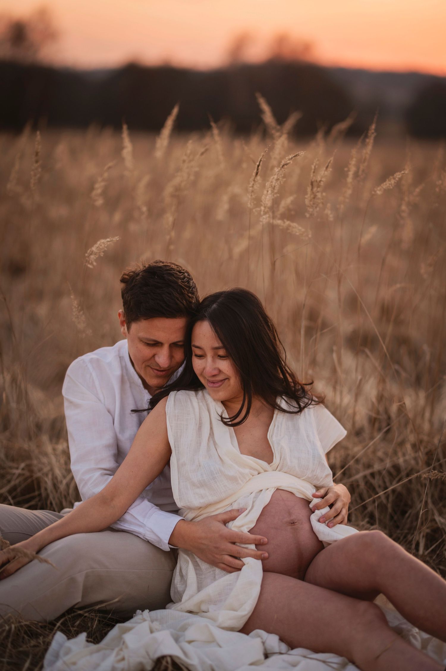 A couple in white outfits sits intimately in a wheat field during golden hour sunset.