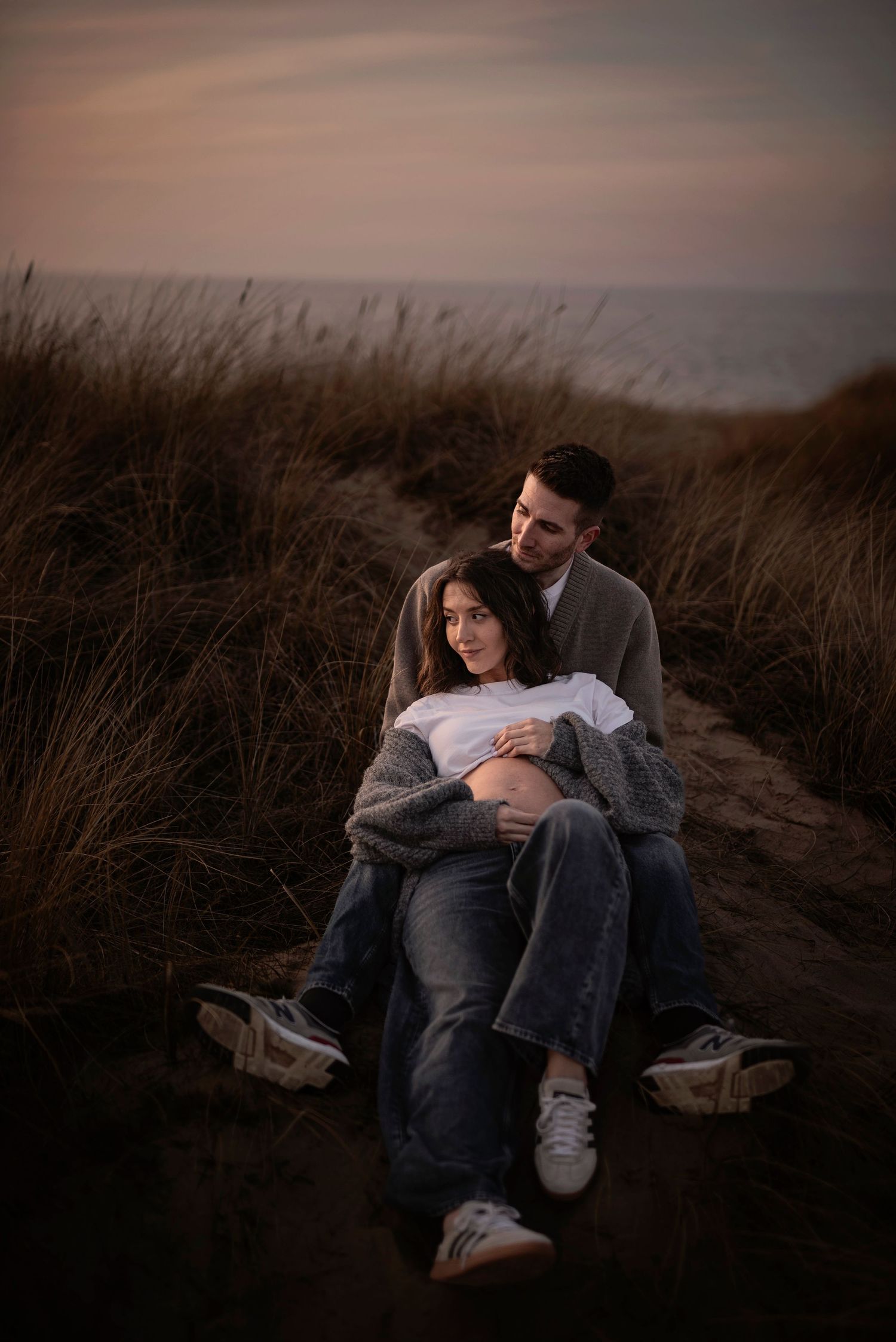 A couple embraces on a coastal path at dusk surrounded by tall grasses and moody skies.