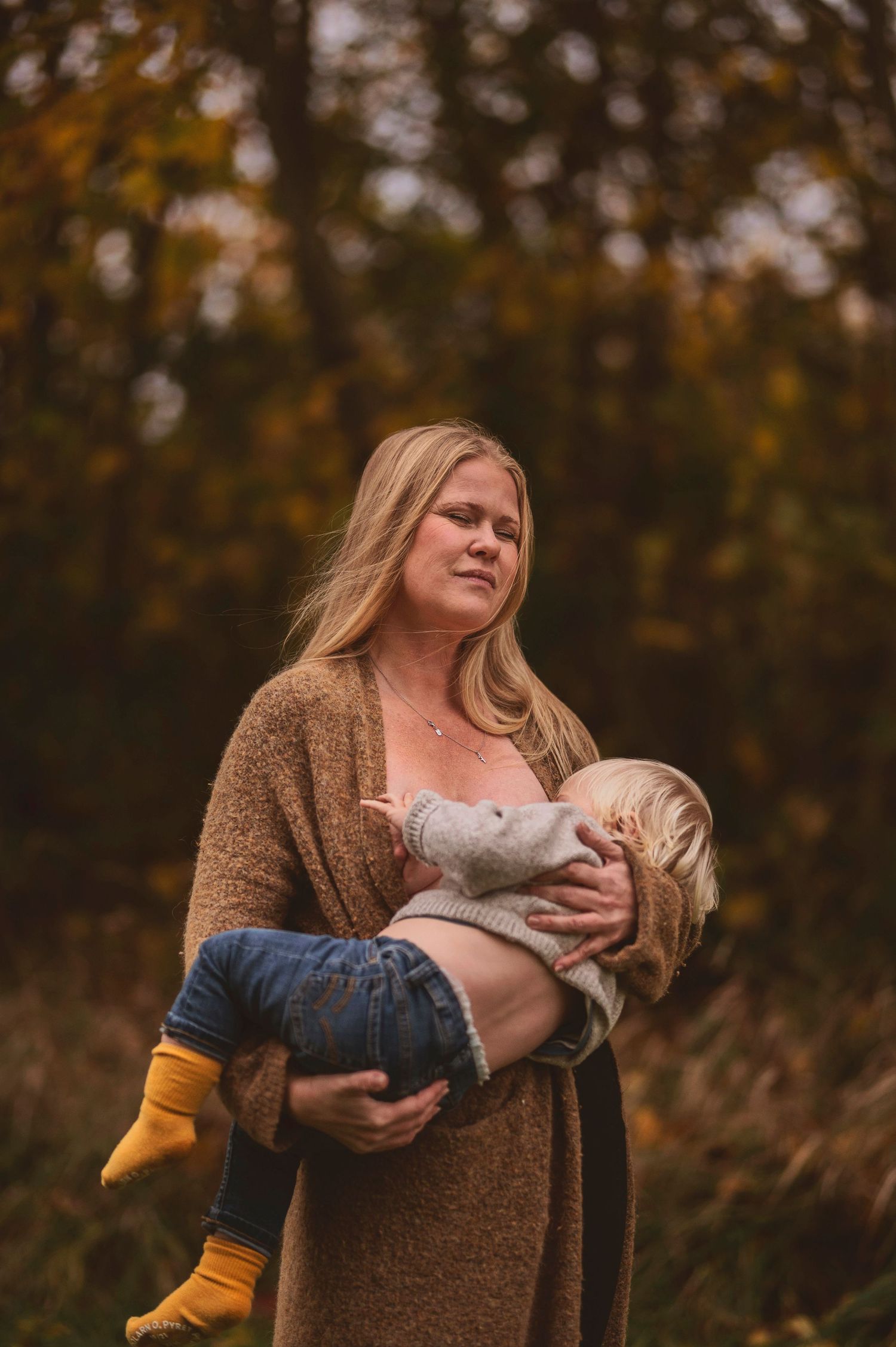 Person in brown cardigan holds small child wearing yellow socks against fall foliage.