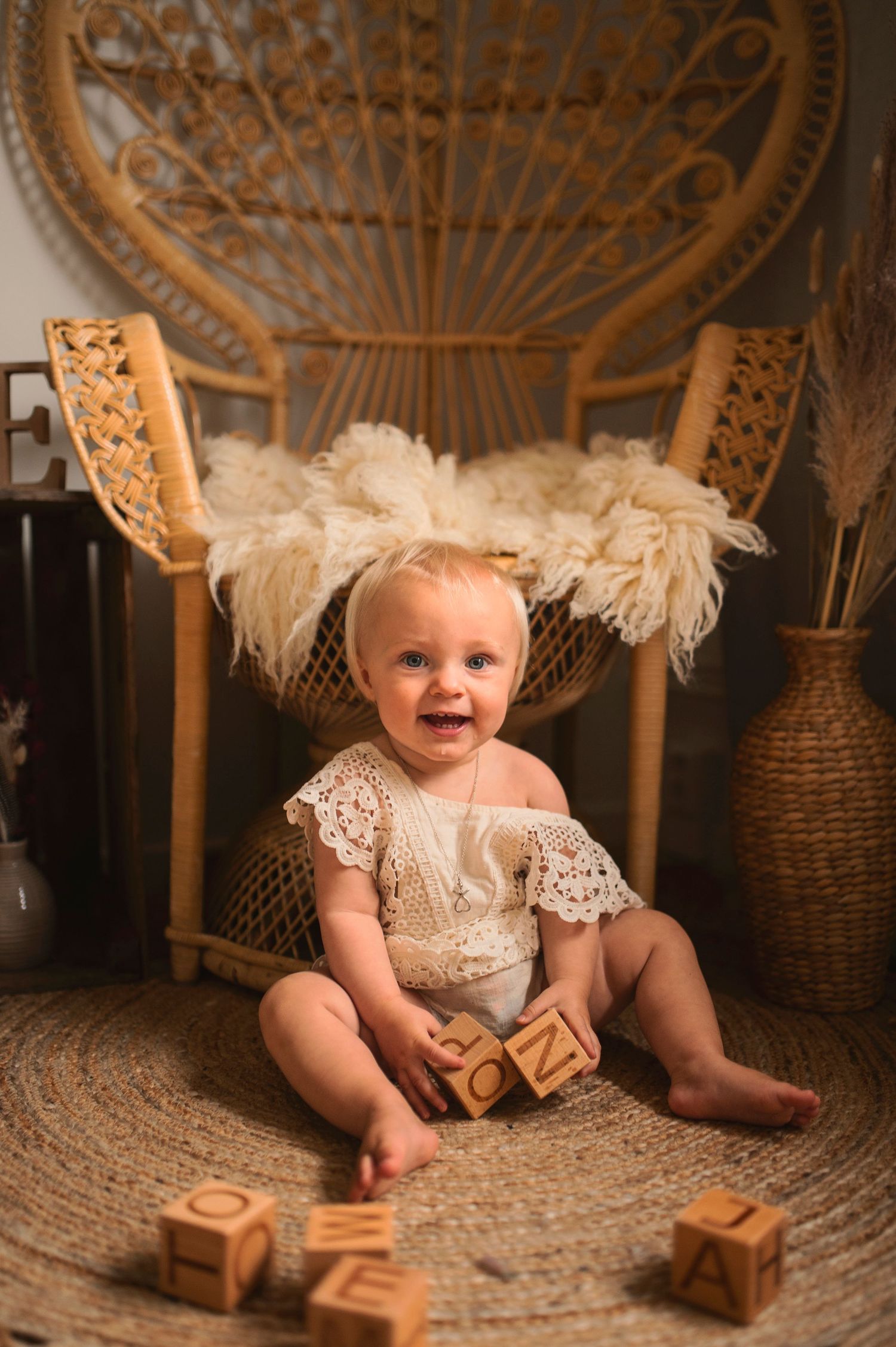Baby in white lace outfit sitting on fluffy rug with wooden blocks in vintage boho setting.