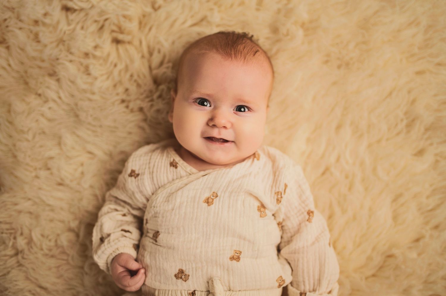 Baby in embroidered cream sweater smiling on plush beige background.