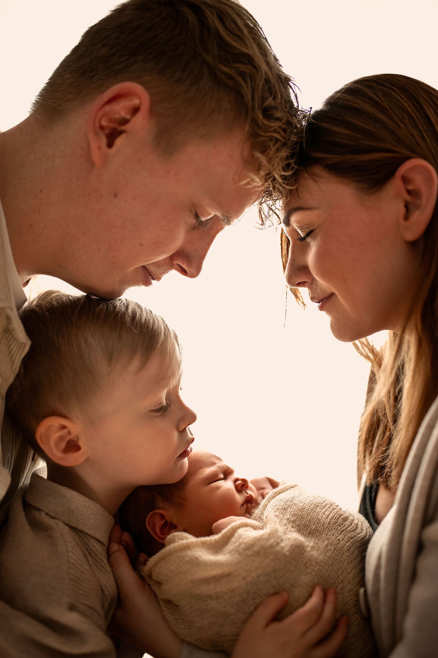 Tender family portrait of parents and toddler gazing lovingly at their newborn baby wrapped in cream knit.