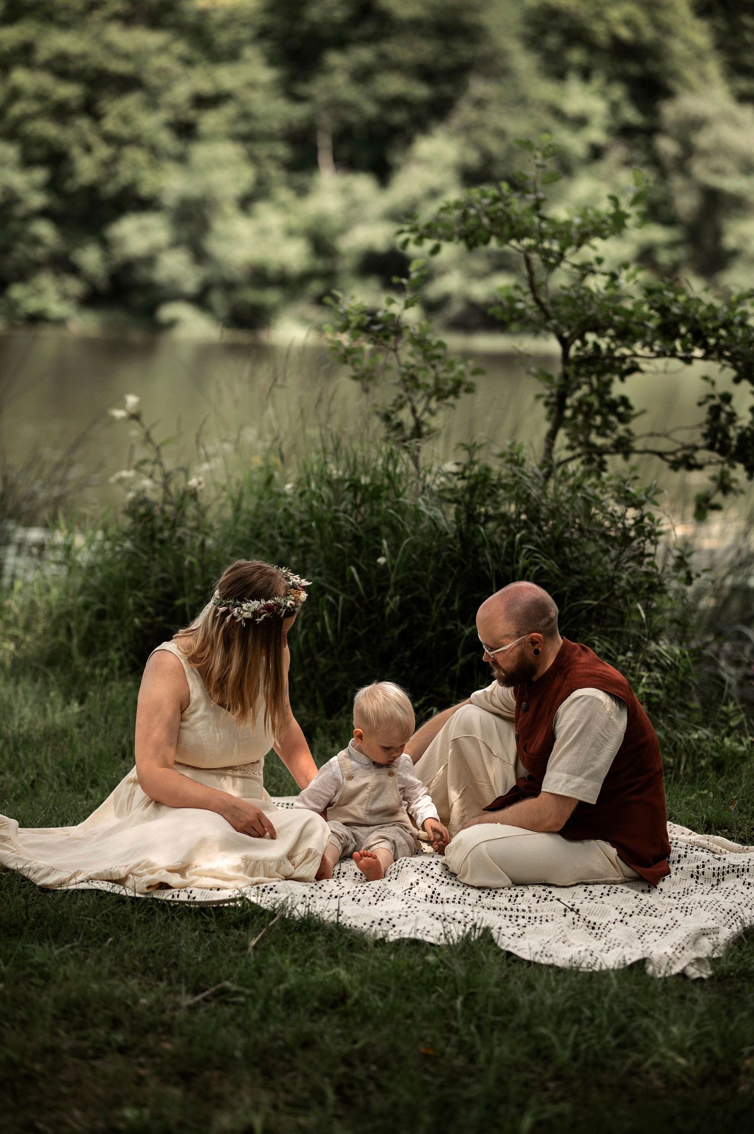 Family with baby sitting on blanket by lake surrounded by lush green nature.