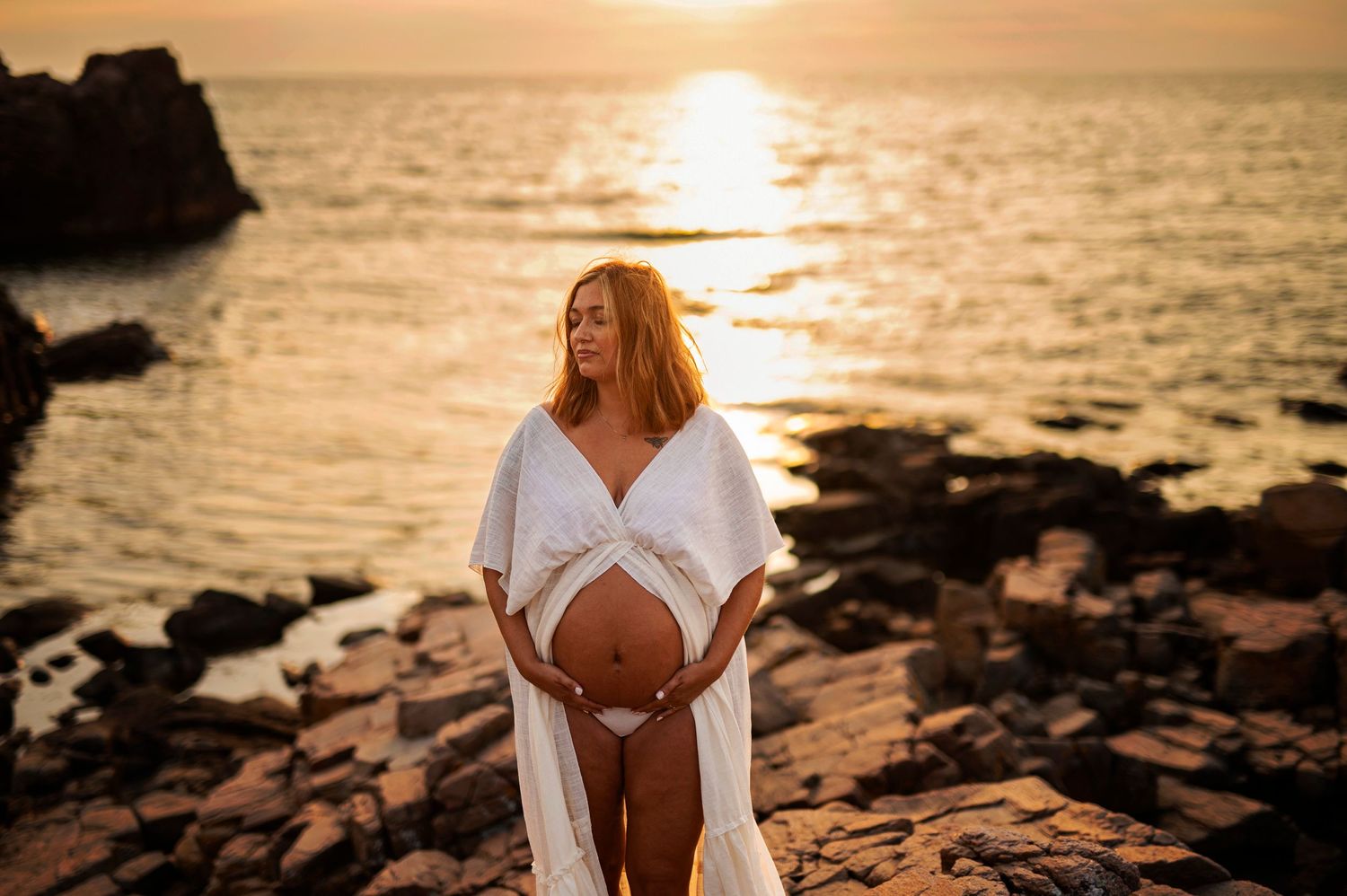 Artistic maternity portrait on rocky beach during golden sunset.
