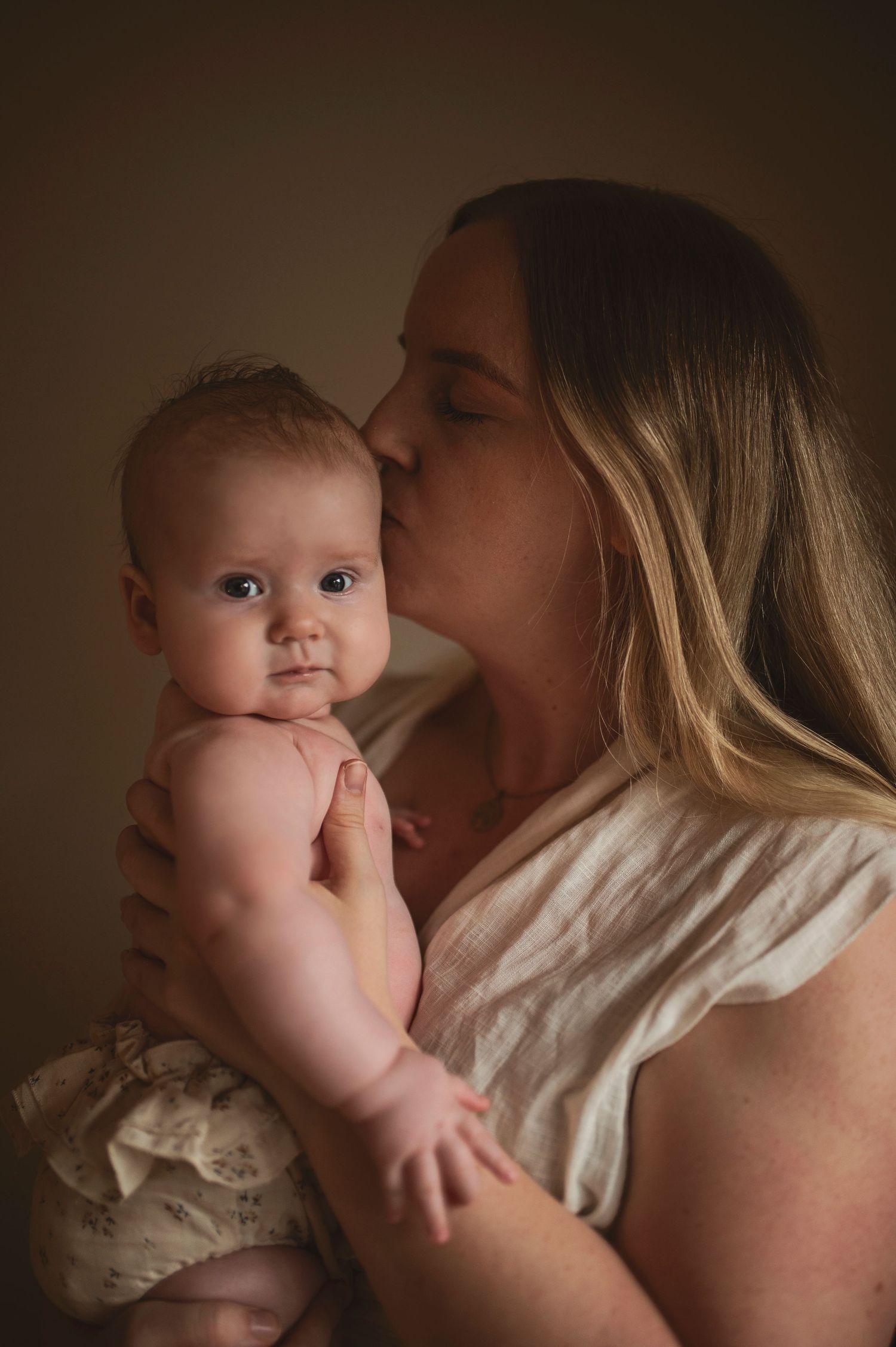 Tender motherhood moment captured in warm tones as parent kisses baby's head.