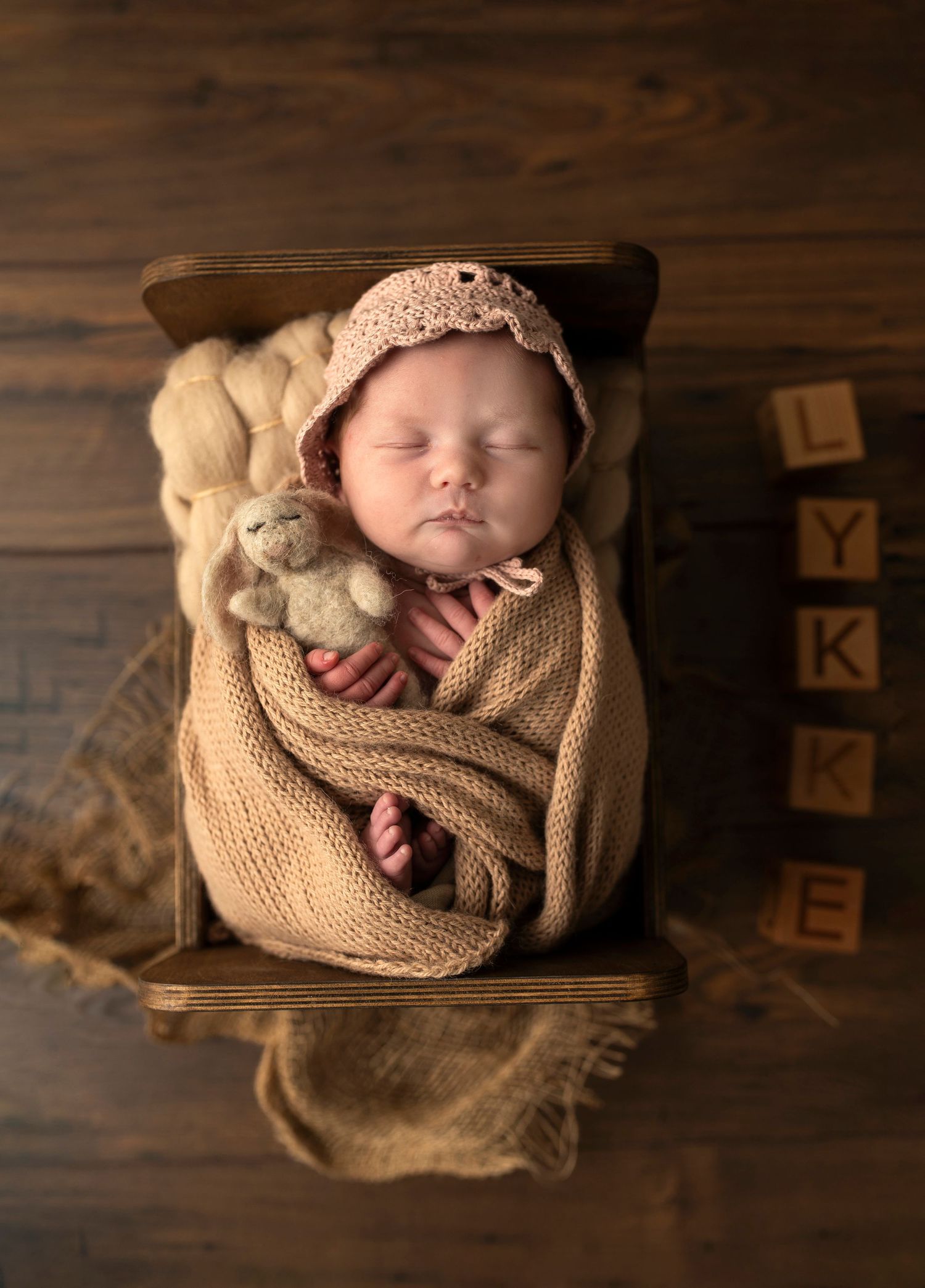 Newborn wrapped in knit blanket sleeps peacefully in wooden box with teddy bear and alphabet blocks.