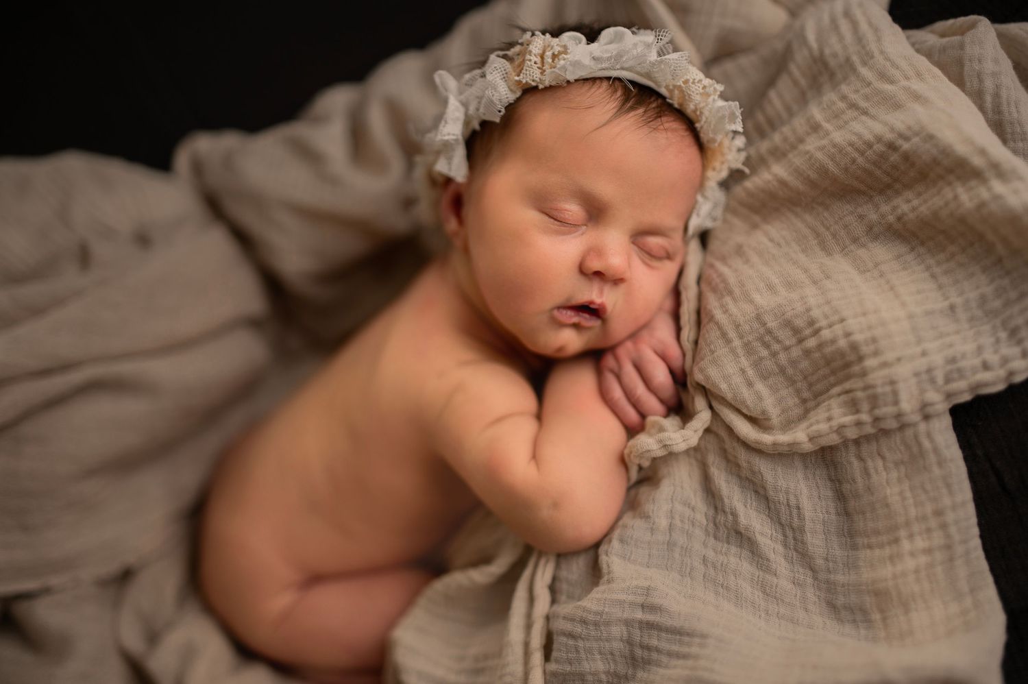 Sleeping newborn wearing a delicate lace bonnet resting on textured beige fabric.