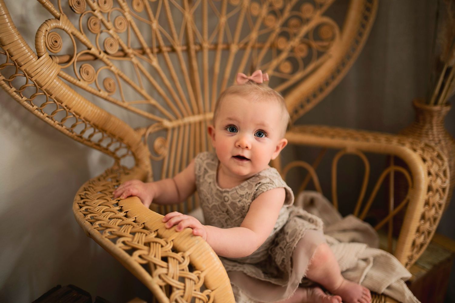 A baby in a lace dress poses in an ornate wicker peacock chair with boho decorations.