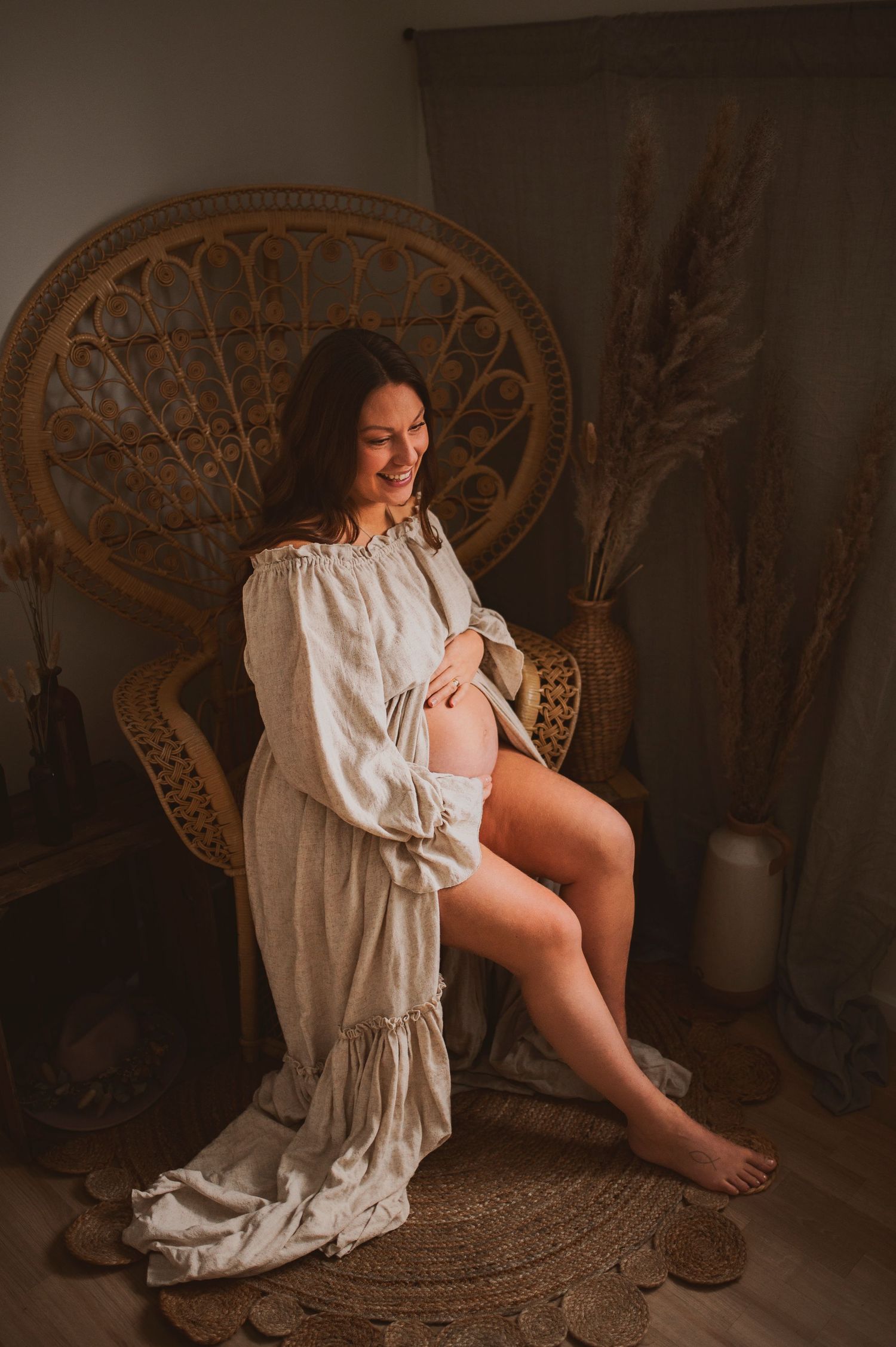 Artistic maternity portrait of expecting mother in white dress seated in woven peacock chair.