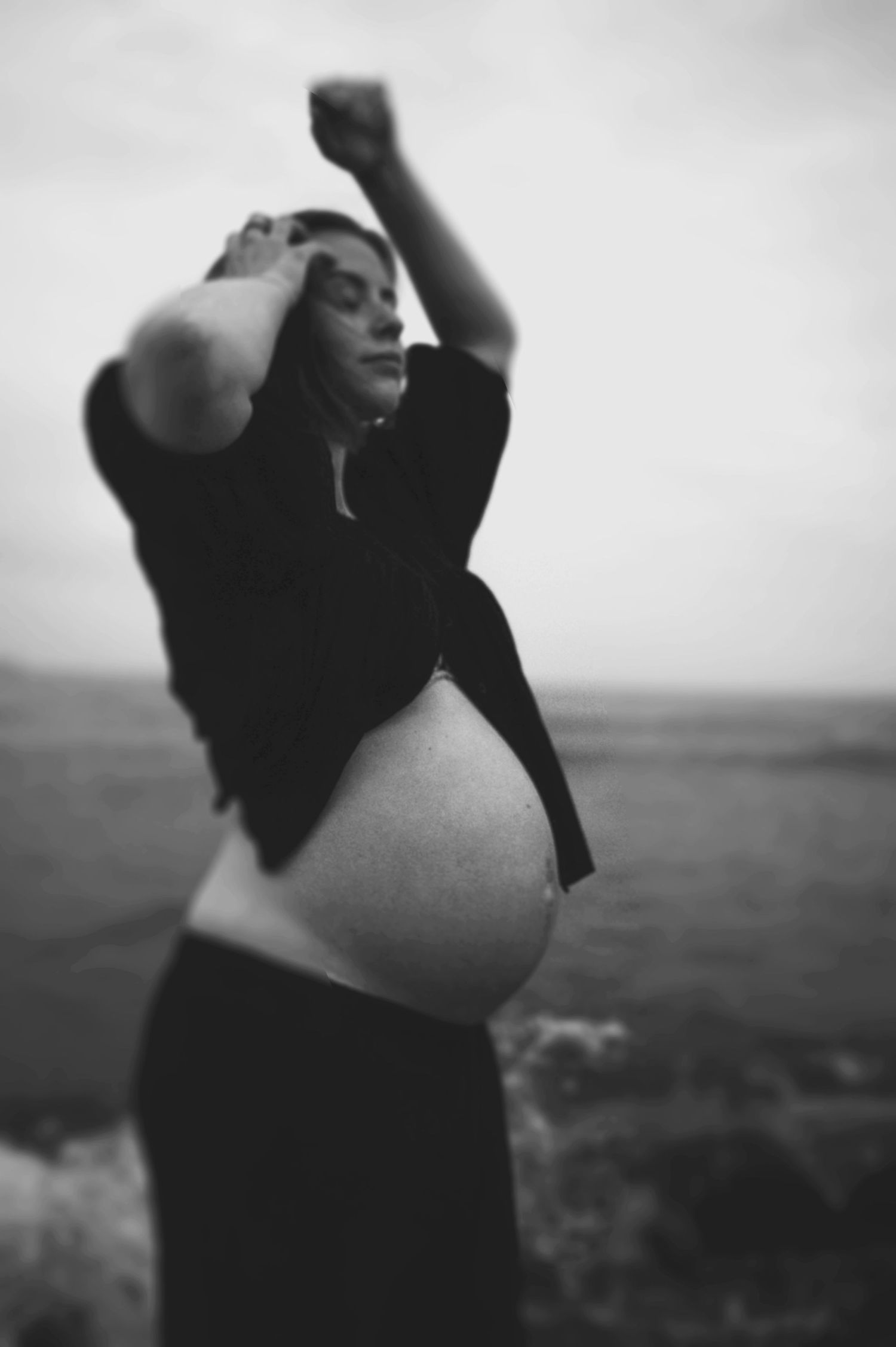 Artistic black and white maternity photo features a pregnant woman in dark clothing against a moody sky.