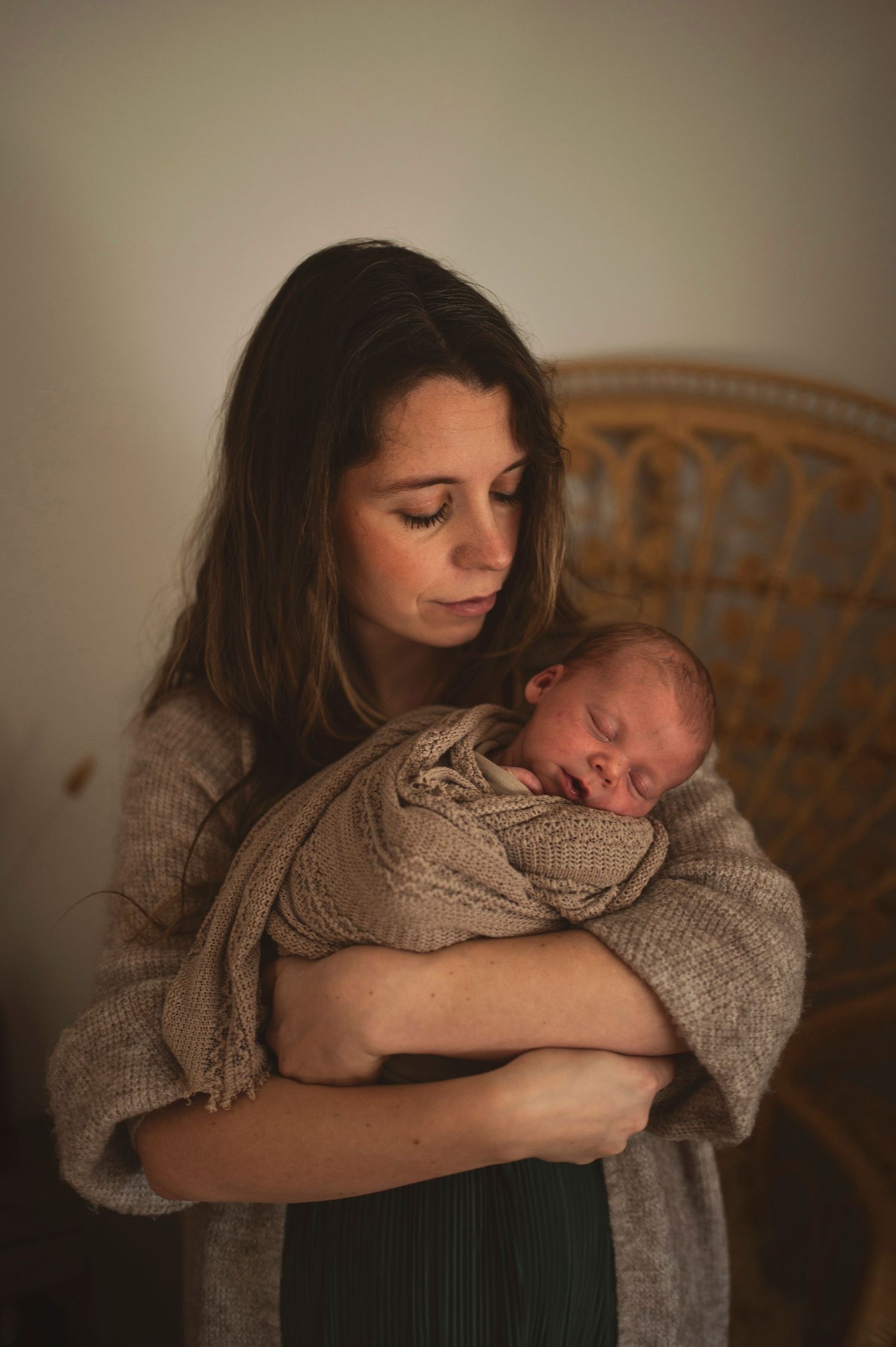 A tender moment captured in warm light as a mother in a beige sweater cradles her sleeping newborn baby.
