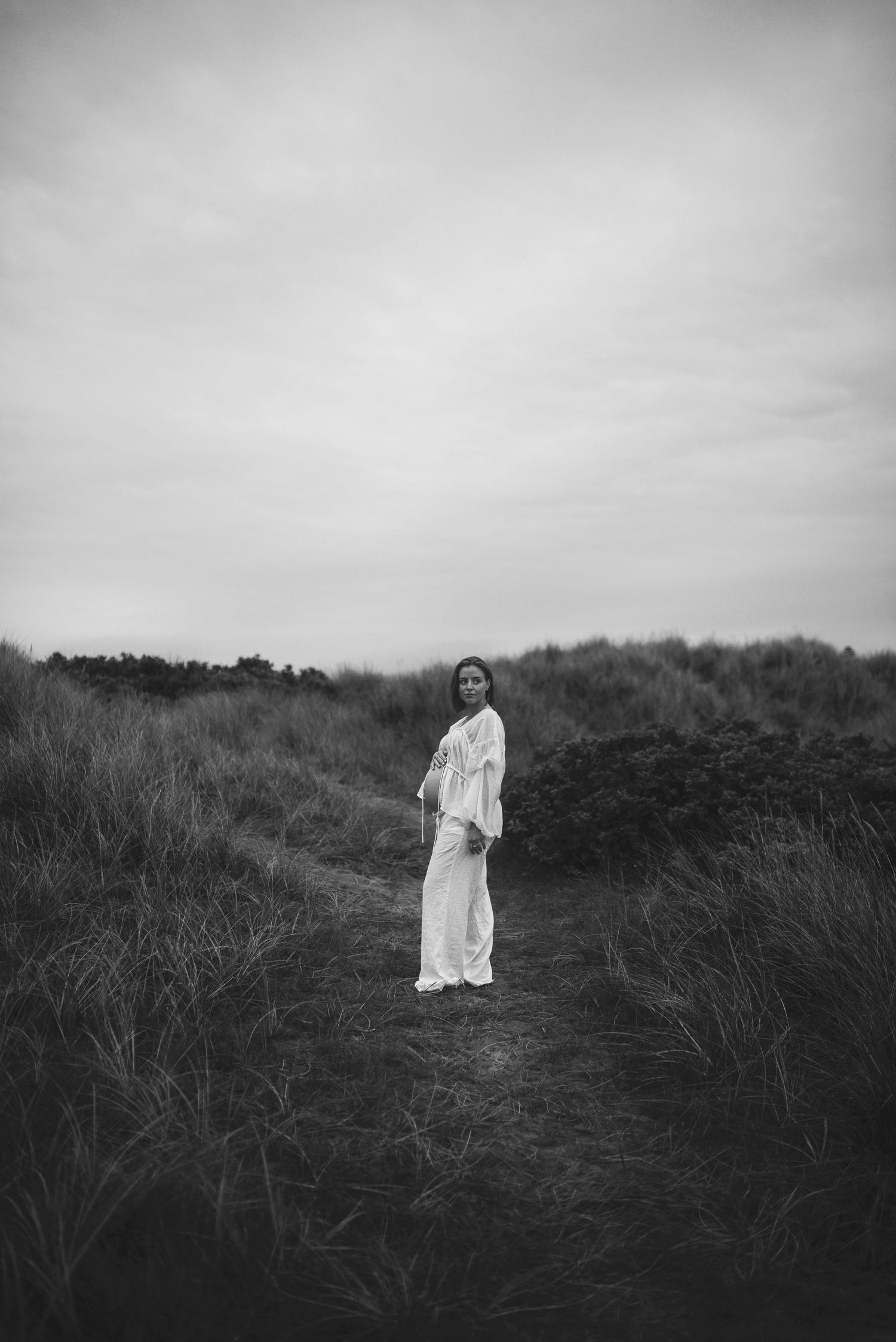 Person in white dress stands on nature trail surrounded by tall grass under moody sky.