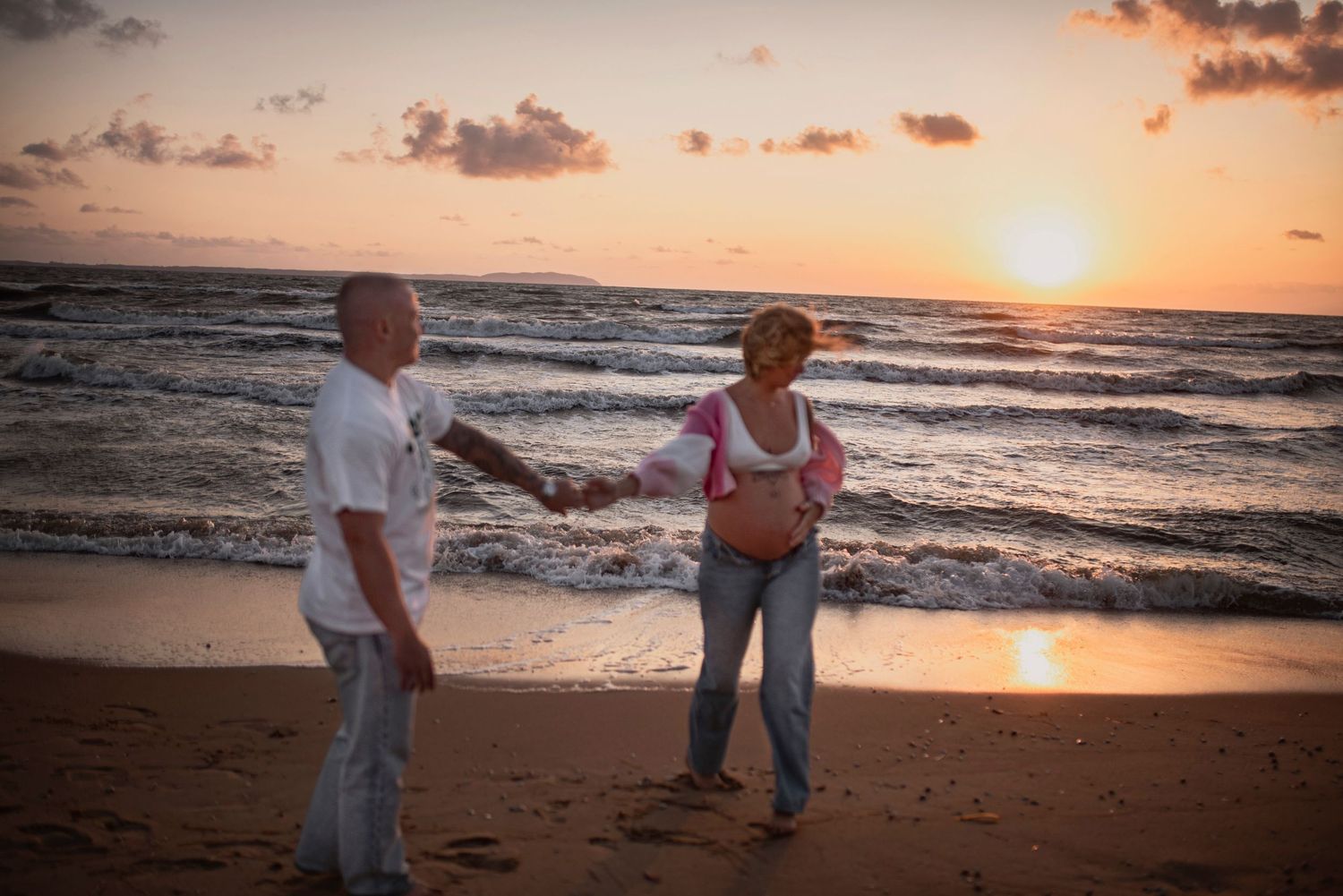 People strolling along sandy beach at sunset with gentle waves and orange skies.