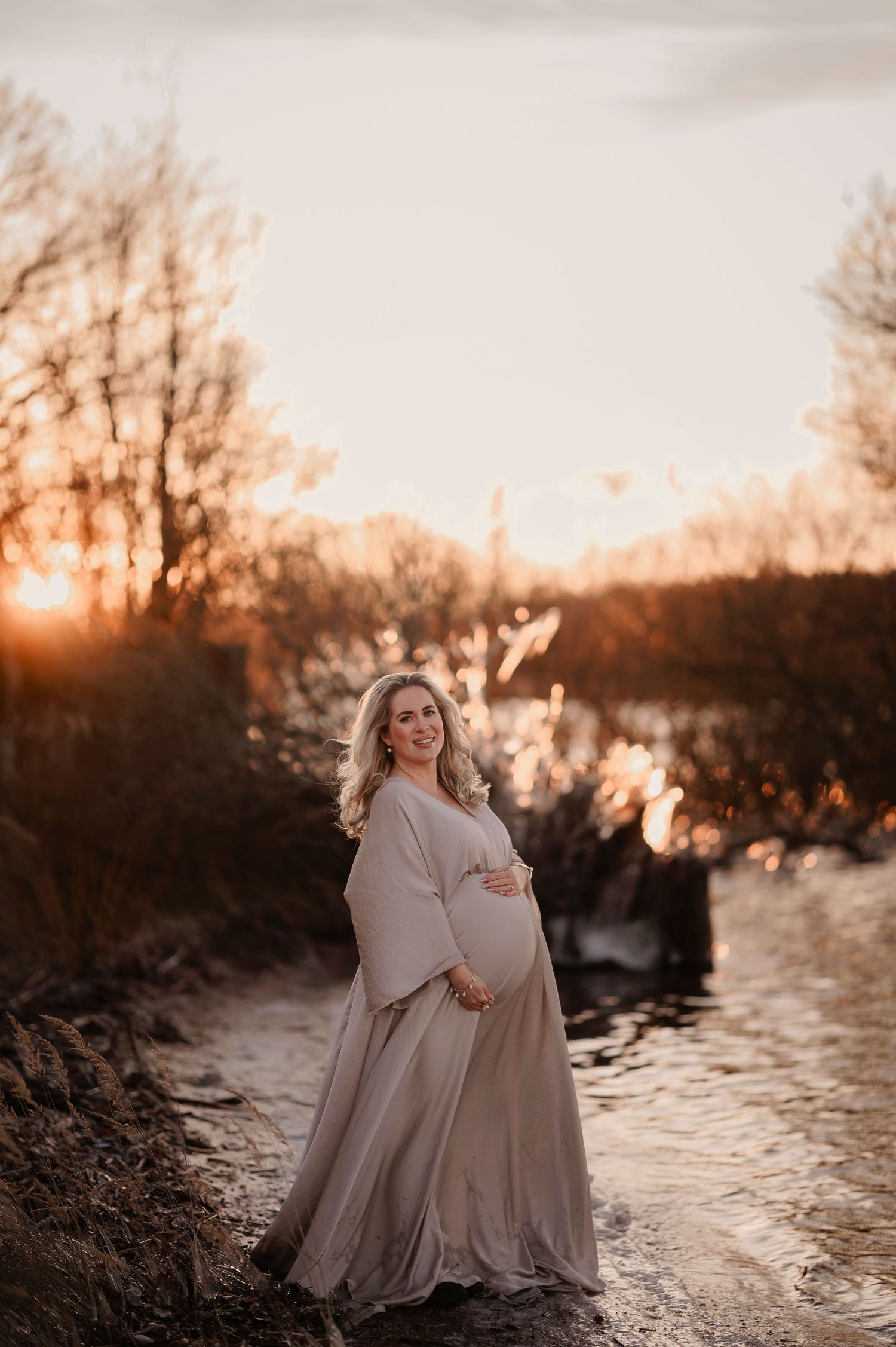 Pregnant woman in flowing beige gown cradling belly at golden sunset by a serene river with bare winter trees.