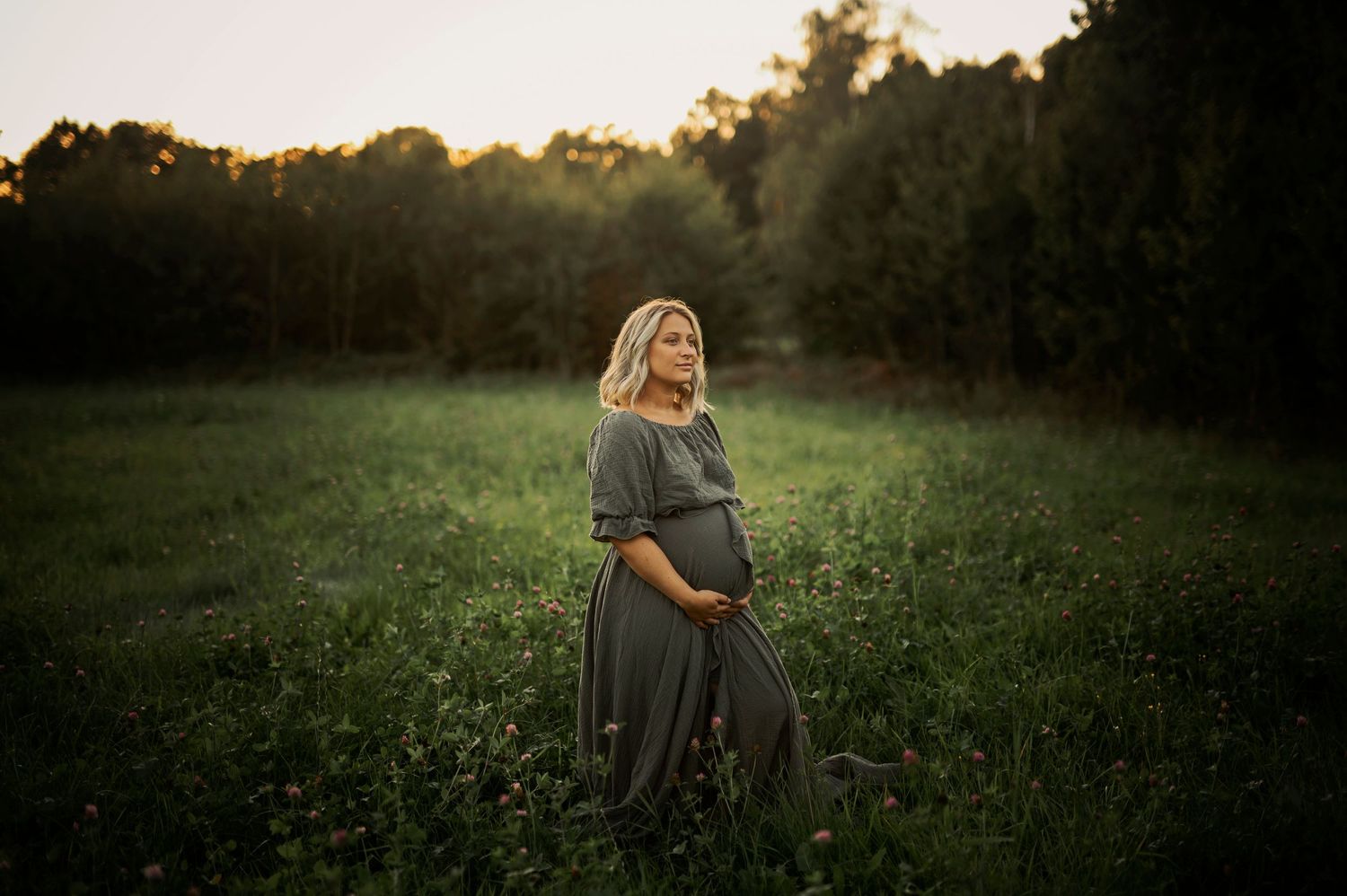 Expectant mother stands in wildflower field wearing gray dress during golden hour.