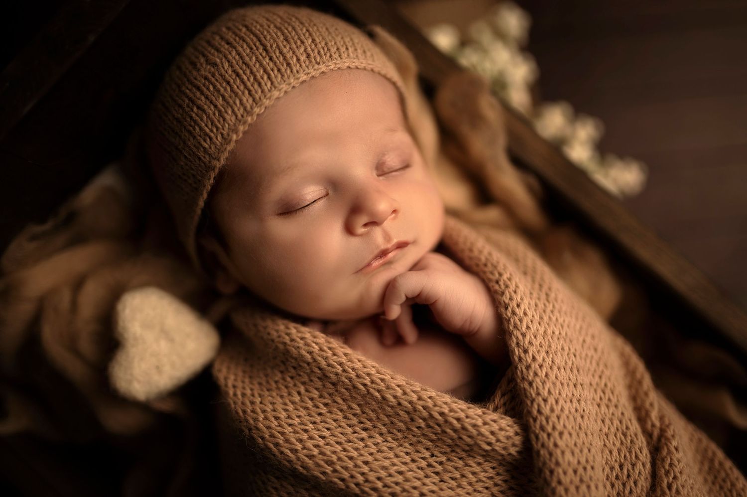Sleeping baby in beige knit wrap and hat against dark background.